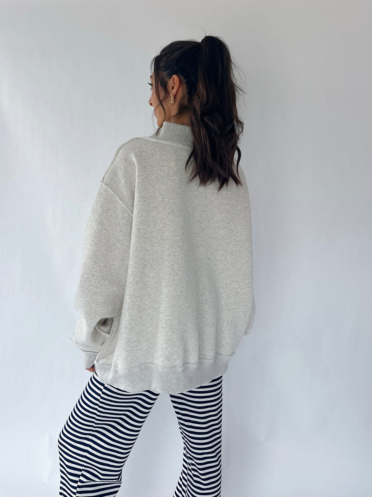 A woman shown from the back wearing an oversized light gray heather sweatshirt and black-and-white striped pants, standing against a plain light gray wall.