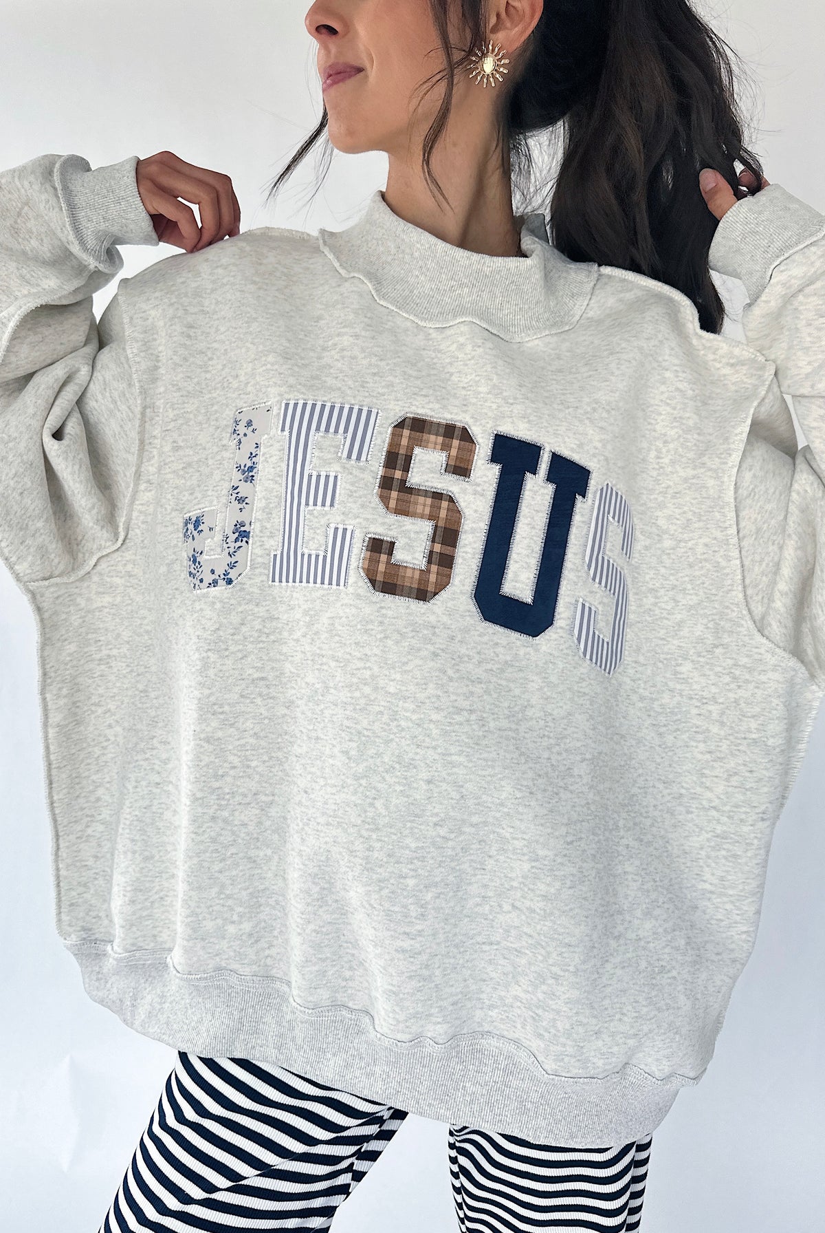 A woman wearing an oversized light gray sweatshirt with 'JESUS' printed across the chest in mixed patterned lettering, paired with black and white striped pants.