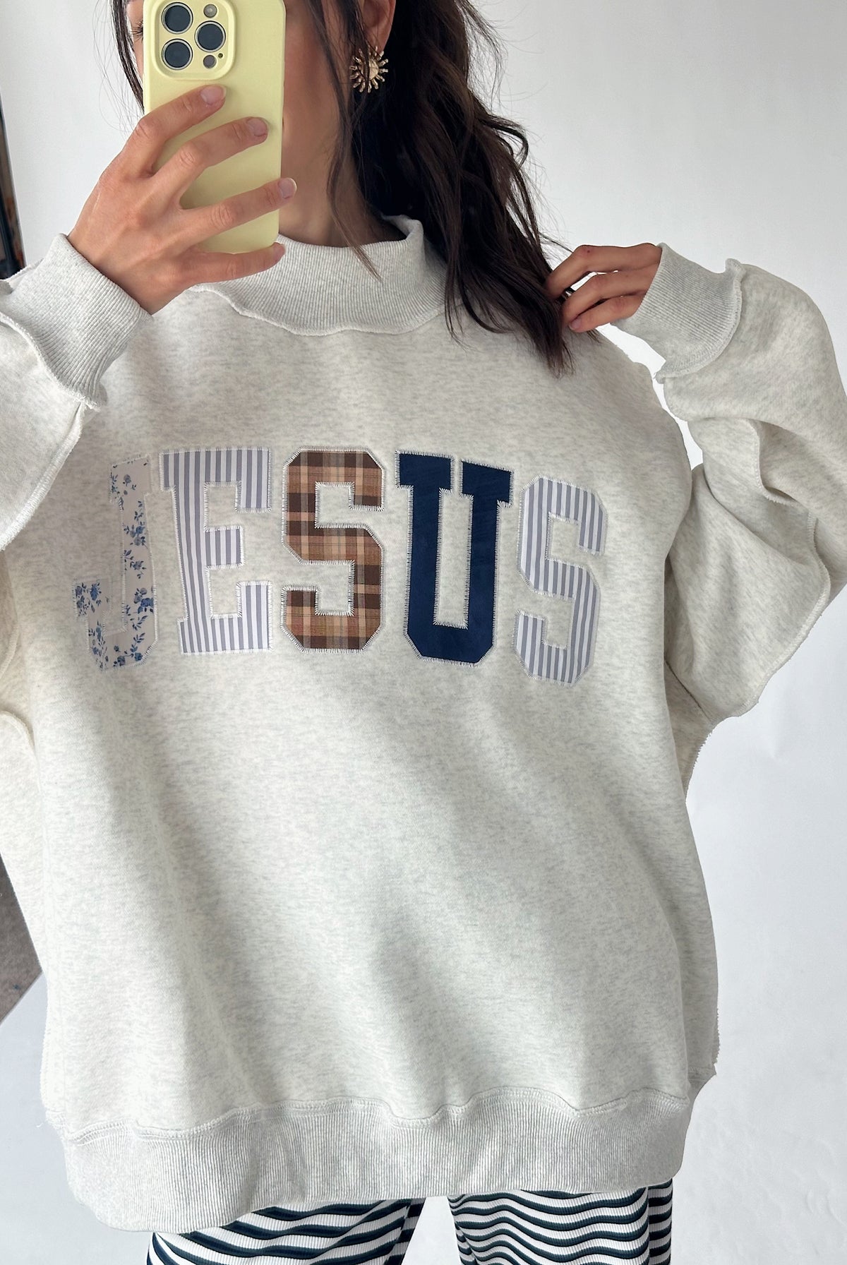 A person wearing a light heather gray sweatshirt with 'JESUS' spelled out across the chest in striped, brown, and navy lettering, holding a phone to take a mirror selfie.