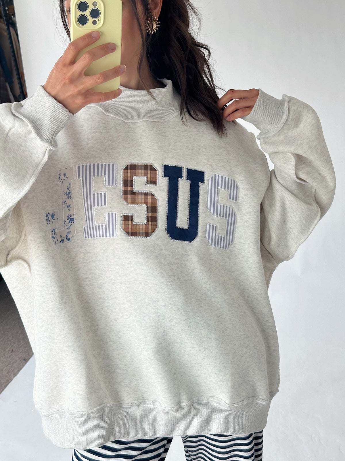 A person wearing a light heather gray sweatshirt with 'JESUS' spelled out across the chest in striped, brown, and navy lettering, holding a phone to take a mirror selfie.