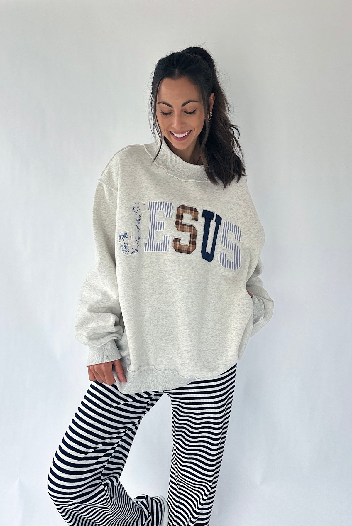 A woman modeling an oversized cream heather sweatshirt with 'JESUS' written across the front in blue, brown, and navy lettering, paired with black and white horizontally striped pants, against a plain white studio background.