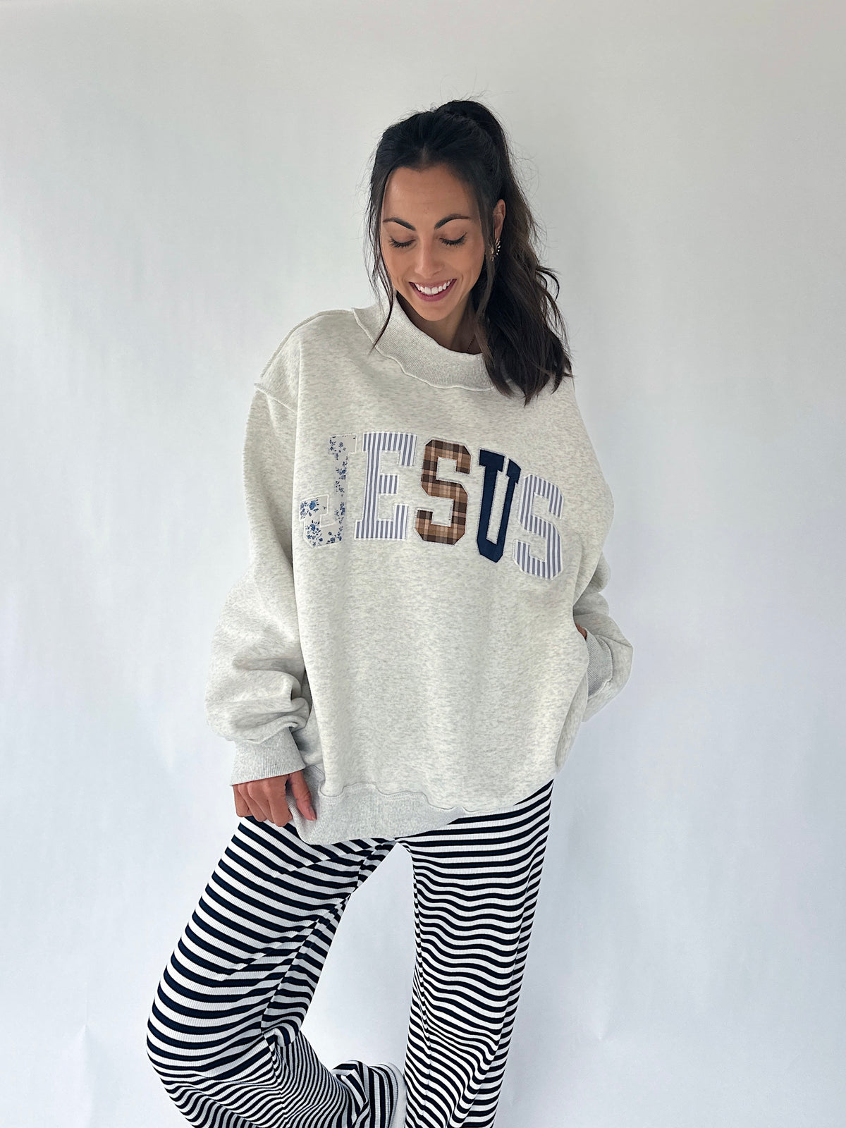 A woman modeling an oversized cream heather sweatshirt with 'JESUS' written across the front in blue, brown, and navy lettering, paired with black and white horizontally striped pants, against a plain white studio background.