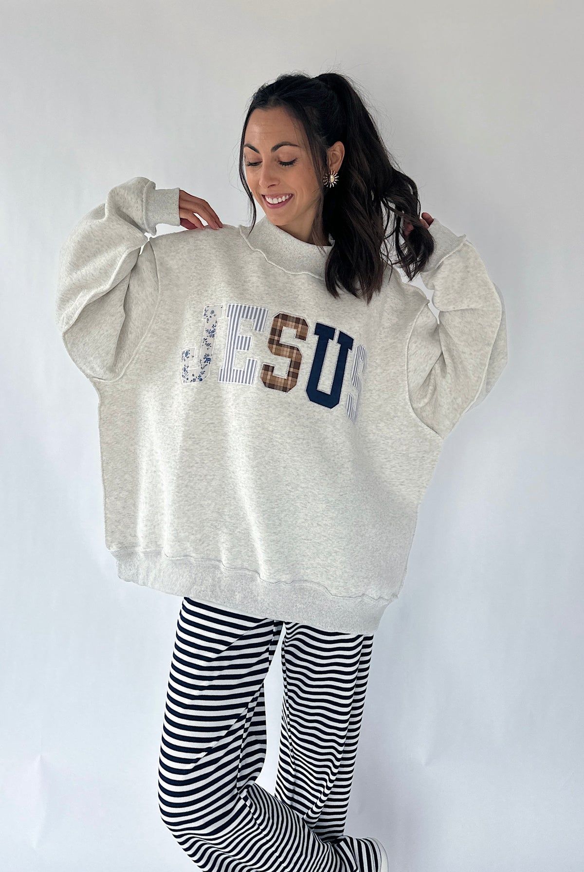 A woman modeling an oversized light heather gray sweatshirt with 'JESUS' printed across the chest in mixed textures, paired with black and white striped pants, against a plain white studio background.