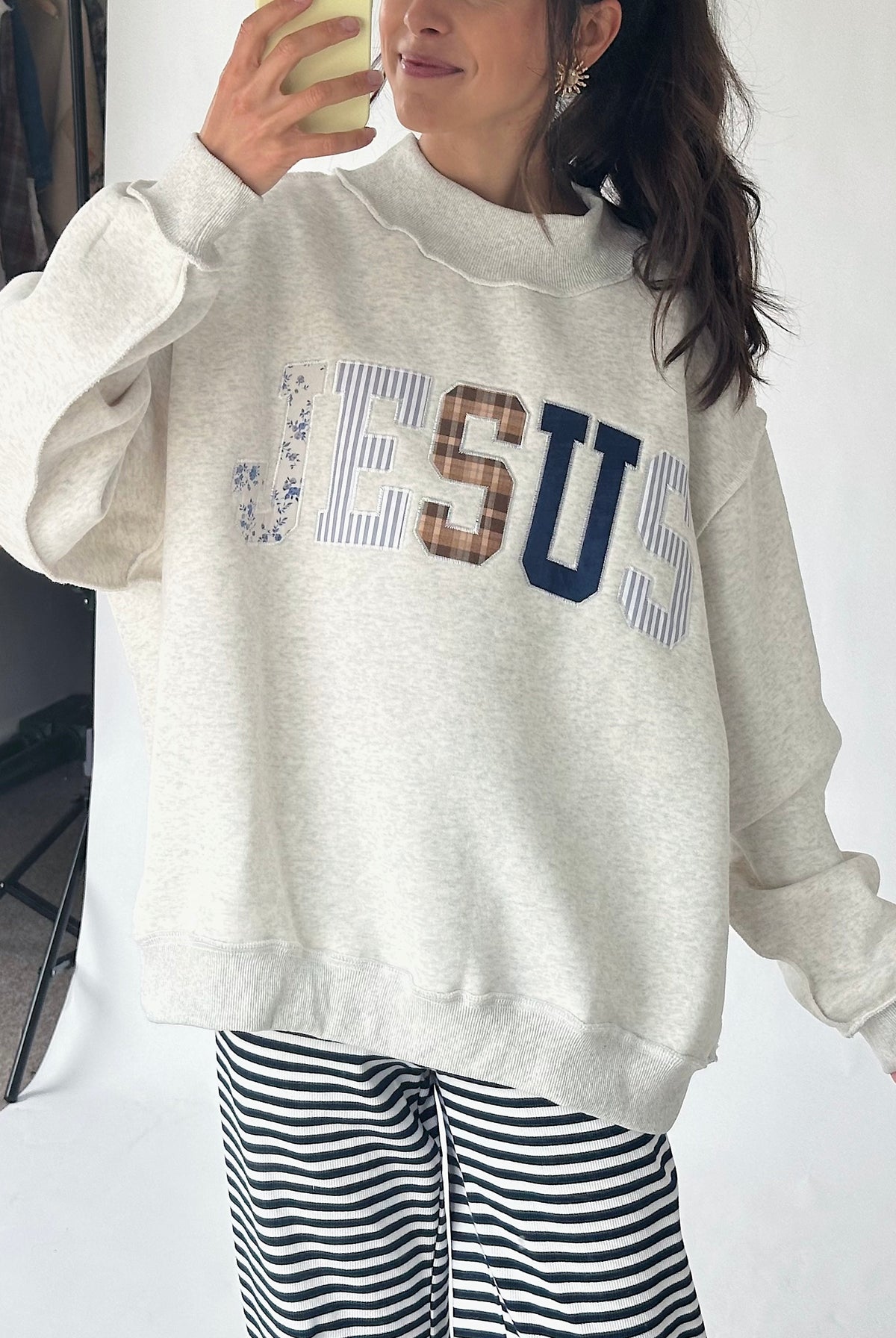A woman wearing an oversized light heather gray sweatshirt with 'JESUS' printed across the chest in blue, brown, and striped lettering, paired with black and white striped pants.