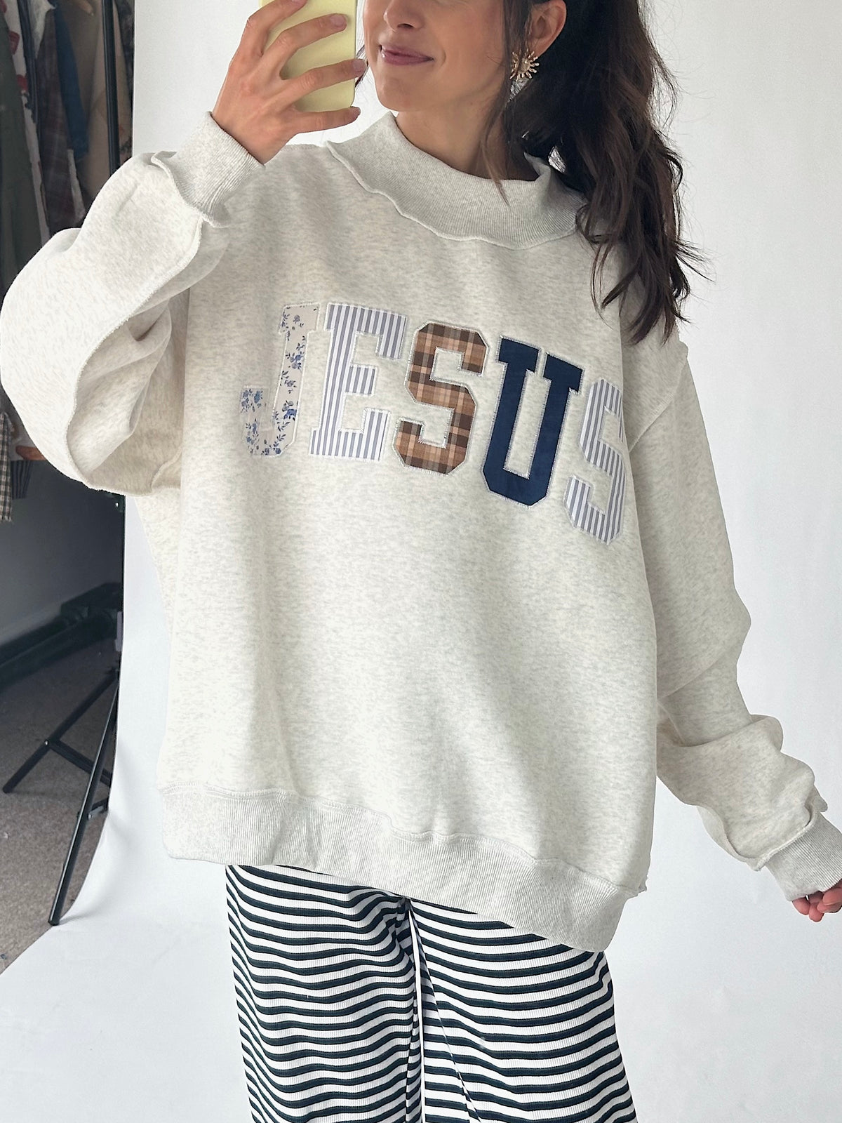 A woman wearing an oversized light heather gray sweatshirt with 'JESUS' printed across the chest in blue, brown, and striped lettering, paired with black and white striped pants.