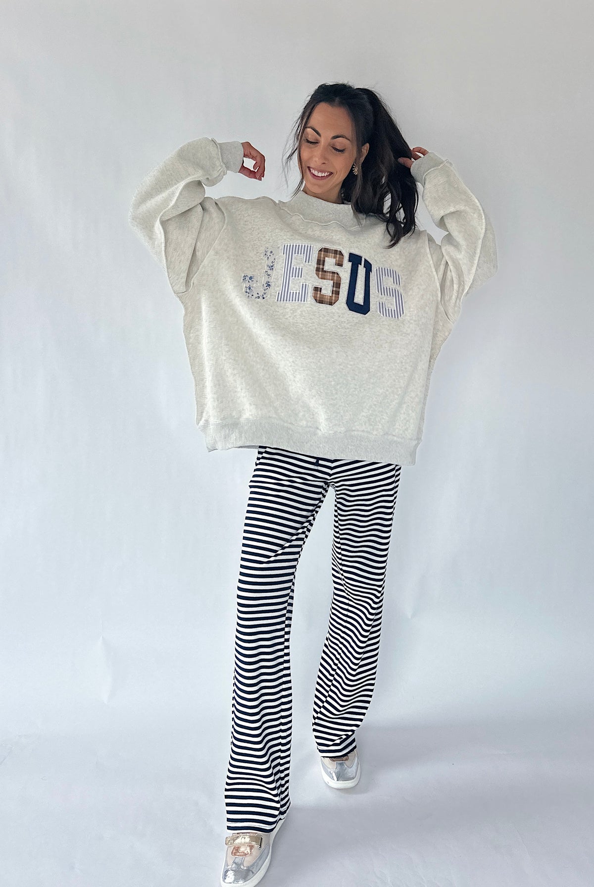 A woman modeling a cream-colored oversized sweatshirt with 'JESUS' written across the chest in blue, brown, and white lettering, paired with black and white horizontally striped pants, against a plain white studio background.