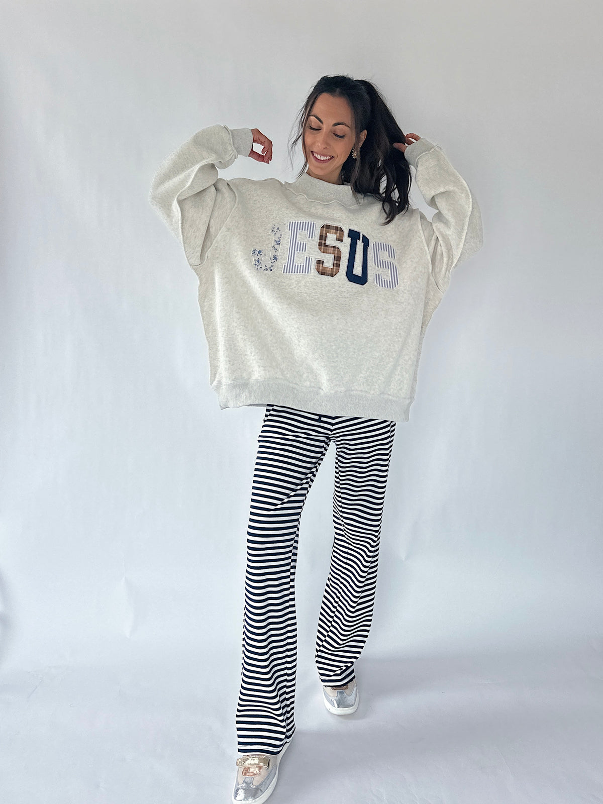 A woman modeling a cream-colored oversized sweatshirt with 'JESUS' written across the chest in blue, brown, and white lettering, paired with black and white horizontally striped pants, against a plain white studio background.
