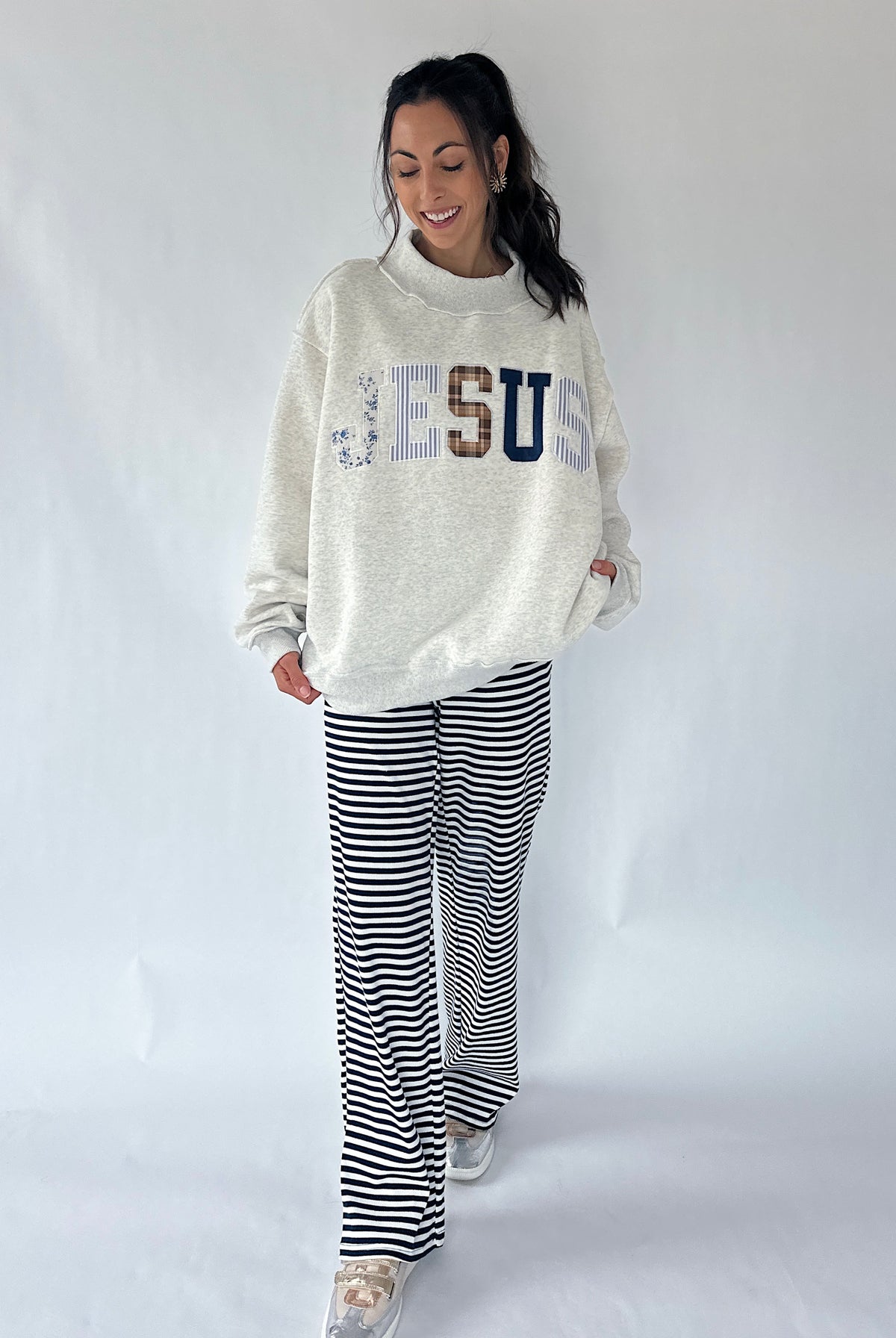 A woman modeling a cream-colored sweatshirt with 'JESUS' spelled out in decorative letters, paired with black and white striped wide-leg pants, against a plain light gray studio background.