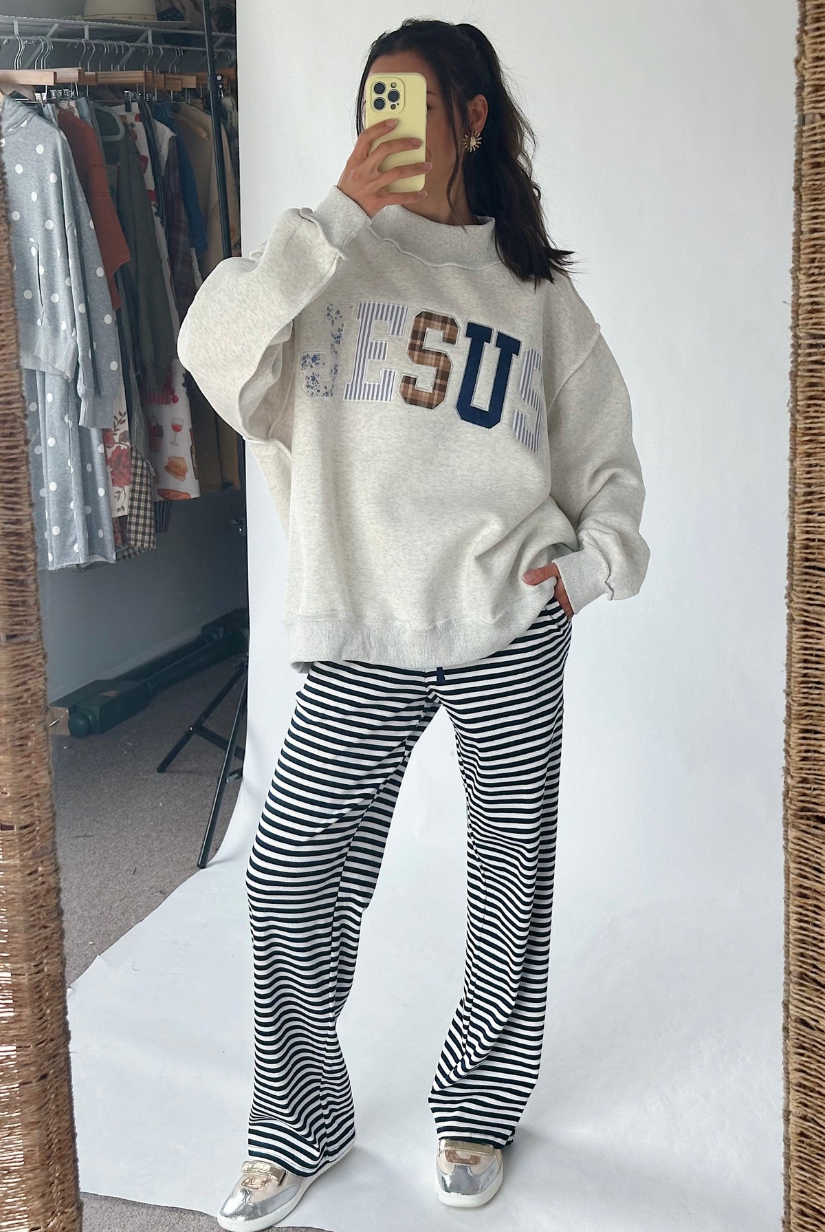 A woman takes a mirror selfie showing a full outfit consisting of a cream-colored sweatshirt with 'JESU' lettering and black-and-white striped pants, in front of a white studio backdrop.