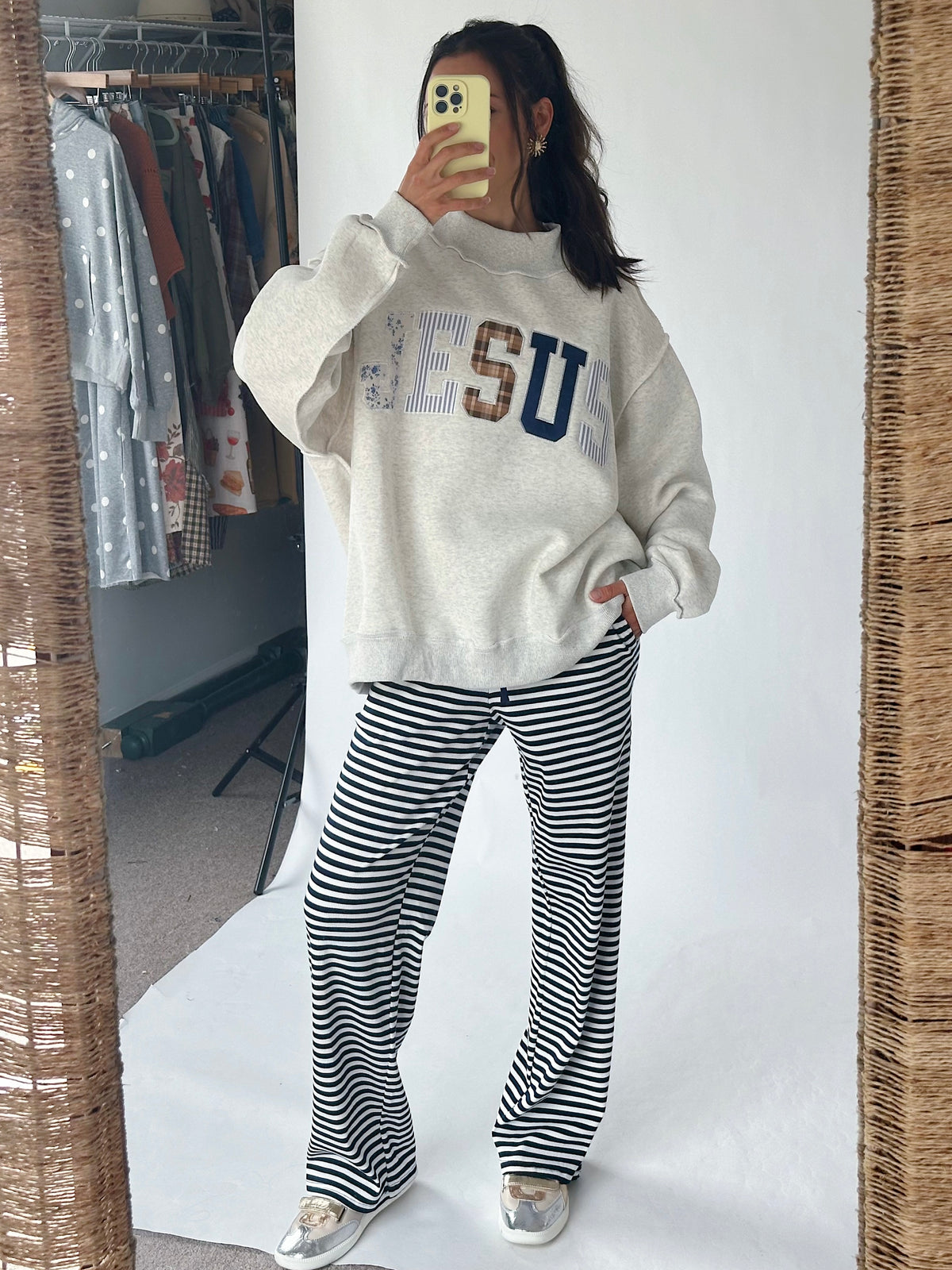 A woman takes a mirror selfie showing a full outfit consisting of a cream-colored sweatshirt with 'JESU' lettering and black-and-white striped pants, in front of a white studio backdrop.