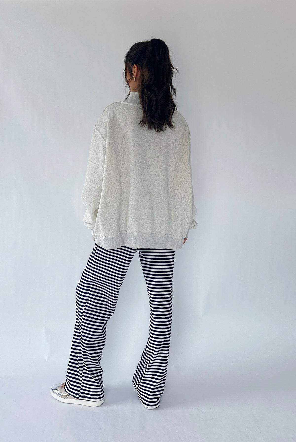 A person standing with their back to the camera, wearing an oversized light gray sweatshirt, black and white horizontally striped flared pants, and white sneakers against a plain white studio background.
