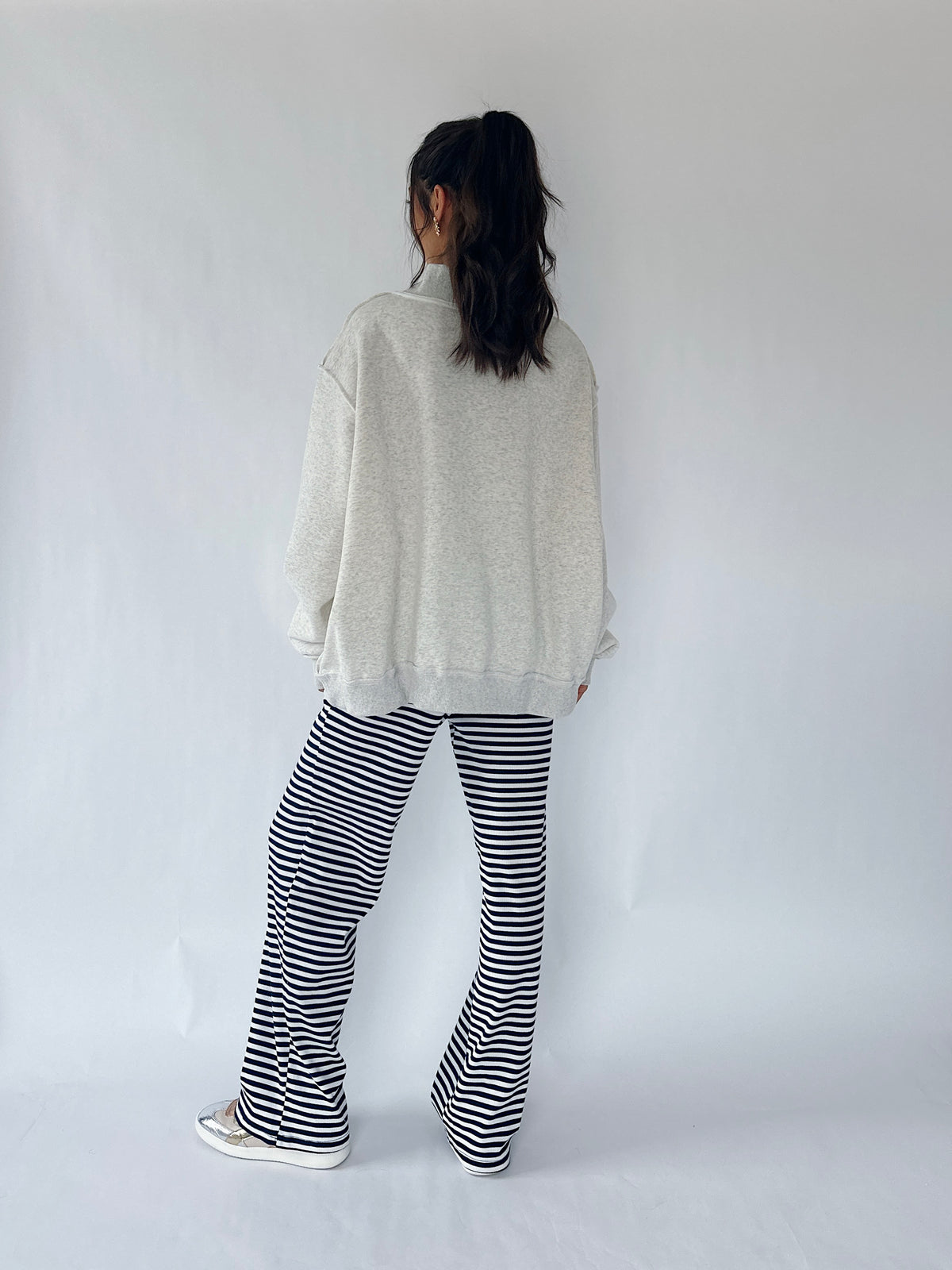 A person standing with their back to the camera, wearing an oversized light gray sweatshirt, black and white horizontally striped flared pants, and white sneakers against a plain white studio background.