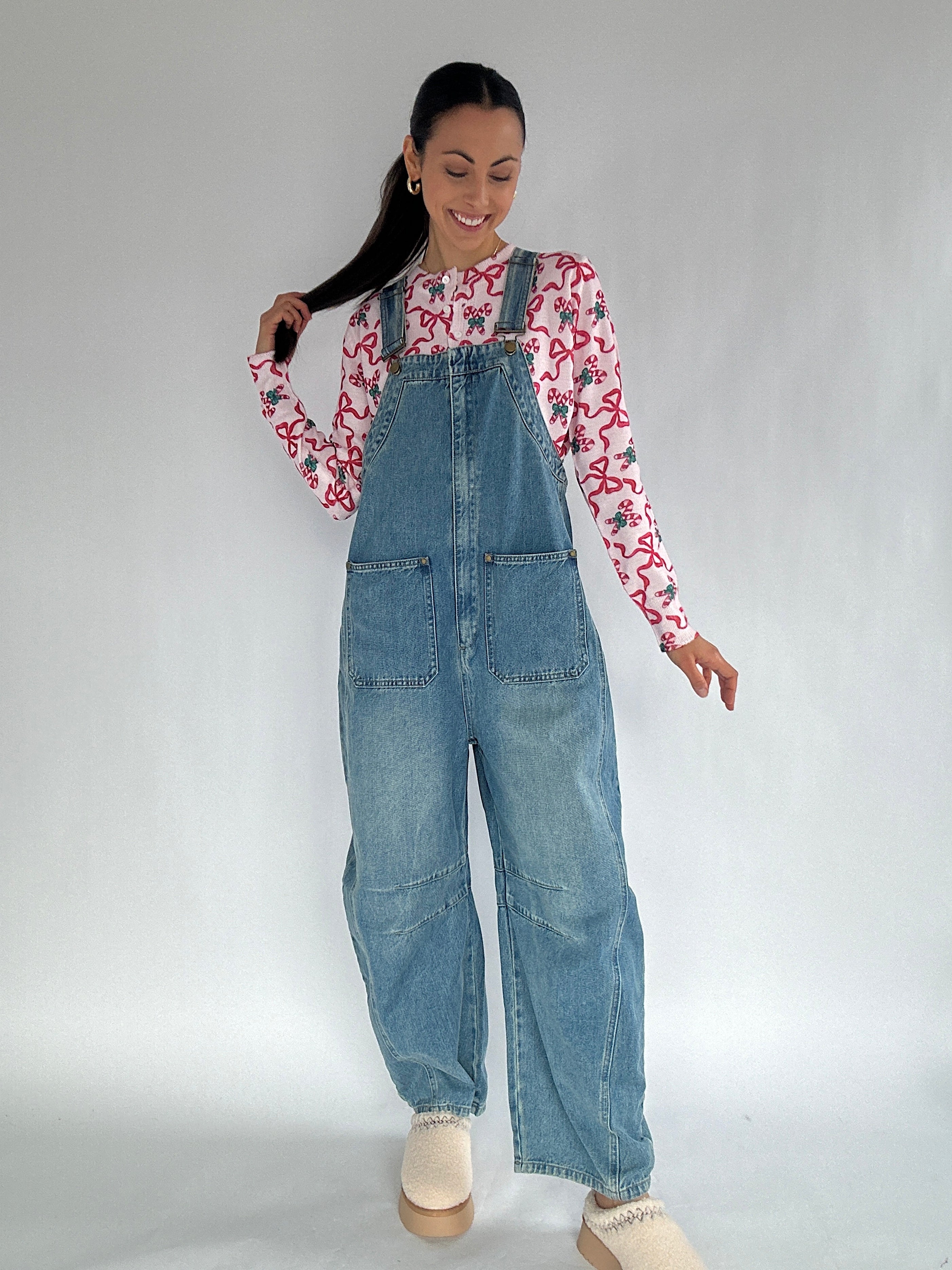Woman wearing a candy cane print long-sleeve cardigan and denim overalls on a plain background