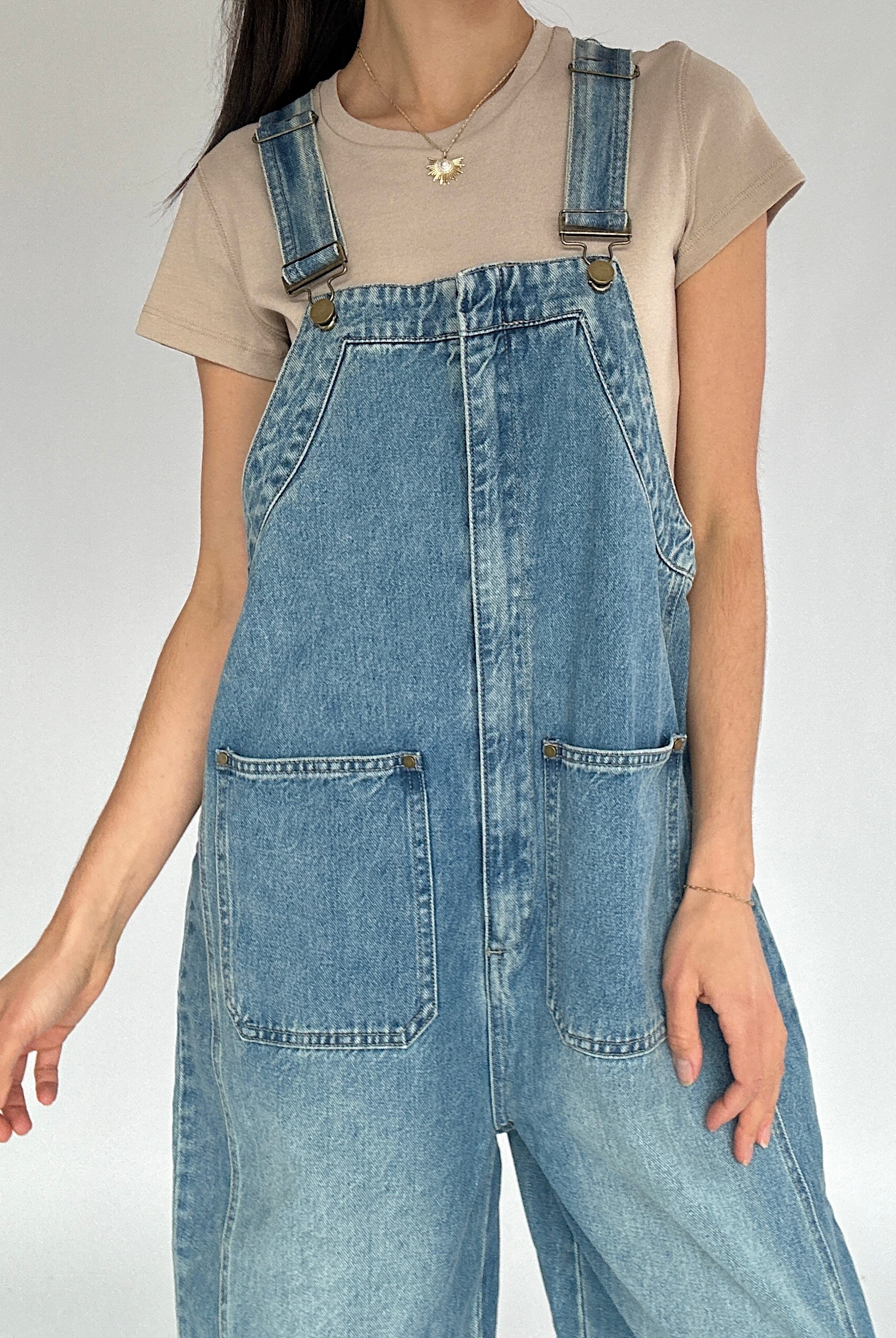 Person wearing blue denim overalls on a plain background showing buckle strap and front pocket details