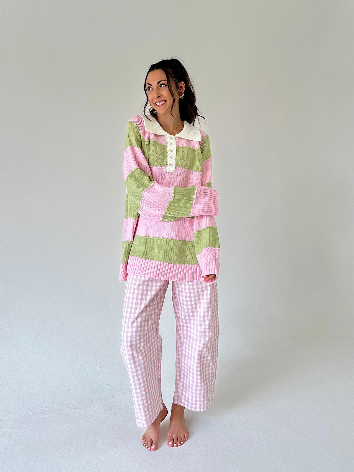 A woman stands barefoot against a plain light gray studio background wearing a pink and sage green striped long-sleeve henley top with a white collar and matching pink-and-white gingham pants.