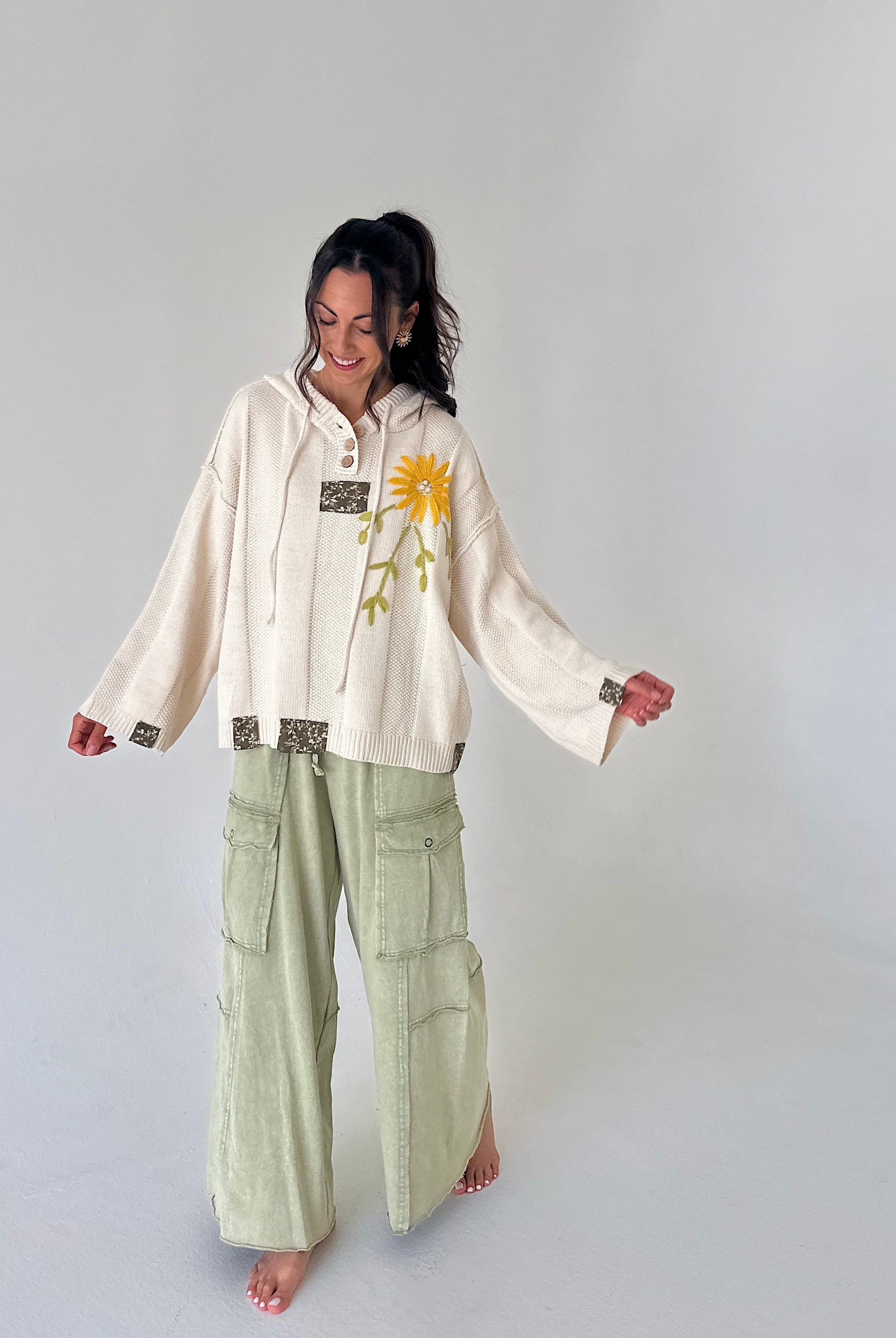 A woman stands barefoot against a light gray studio background, wearing a cream hooded long-sleeve top with a yellow flower graphic and sage green wide-leg cargo pants.
