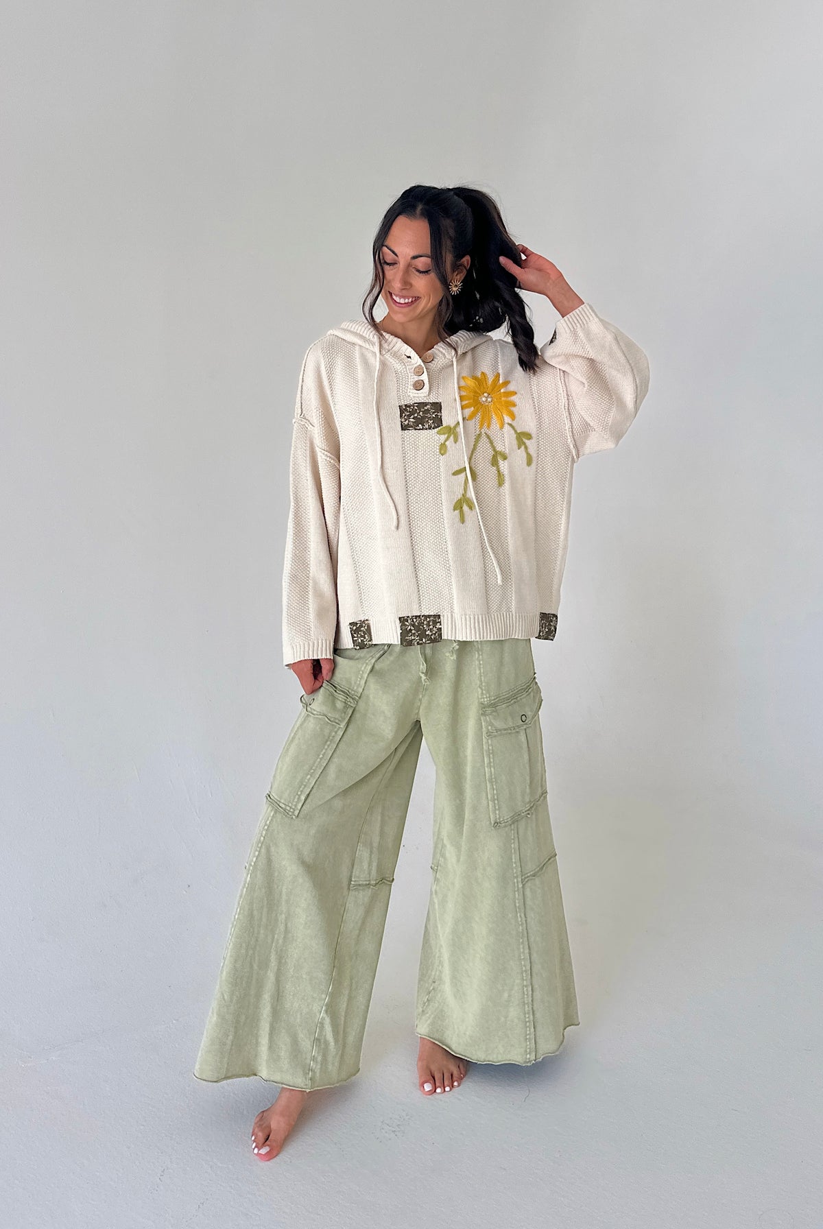 A woman stands barefoot against a light gray studio background, wearing a cream hooded sweatshirt with a yellow flower graphic and green wide-leg cargo pants.