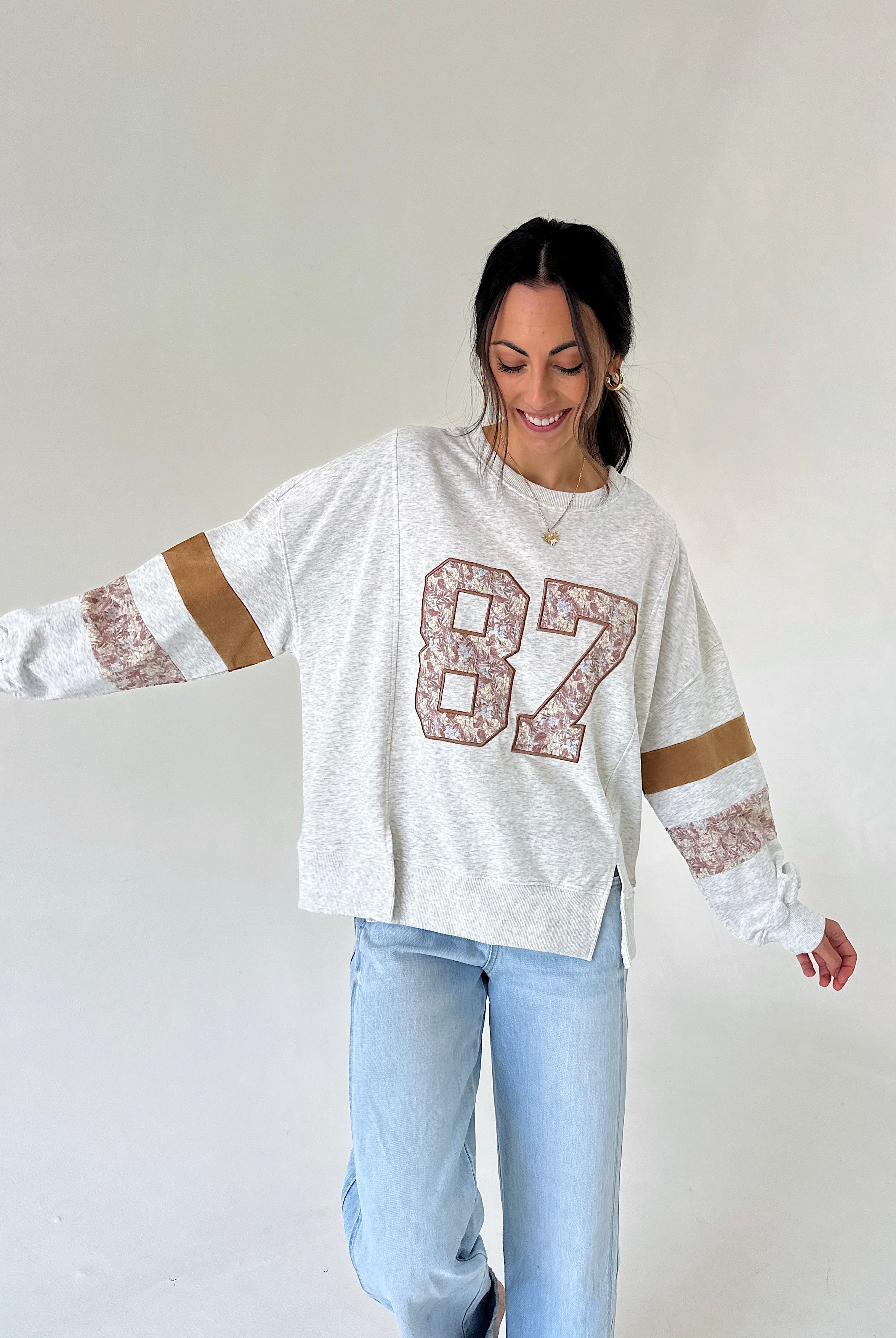 A woman wearing a white heathered long-sleeve sweatshirt with a large '87' graphic on the front, featuring brown and white patterned sleeve stripes, paired with light blue jeans, against a plain off-white background.