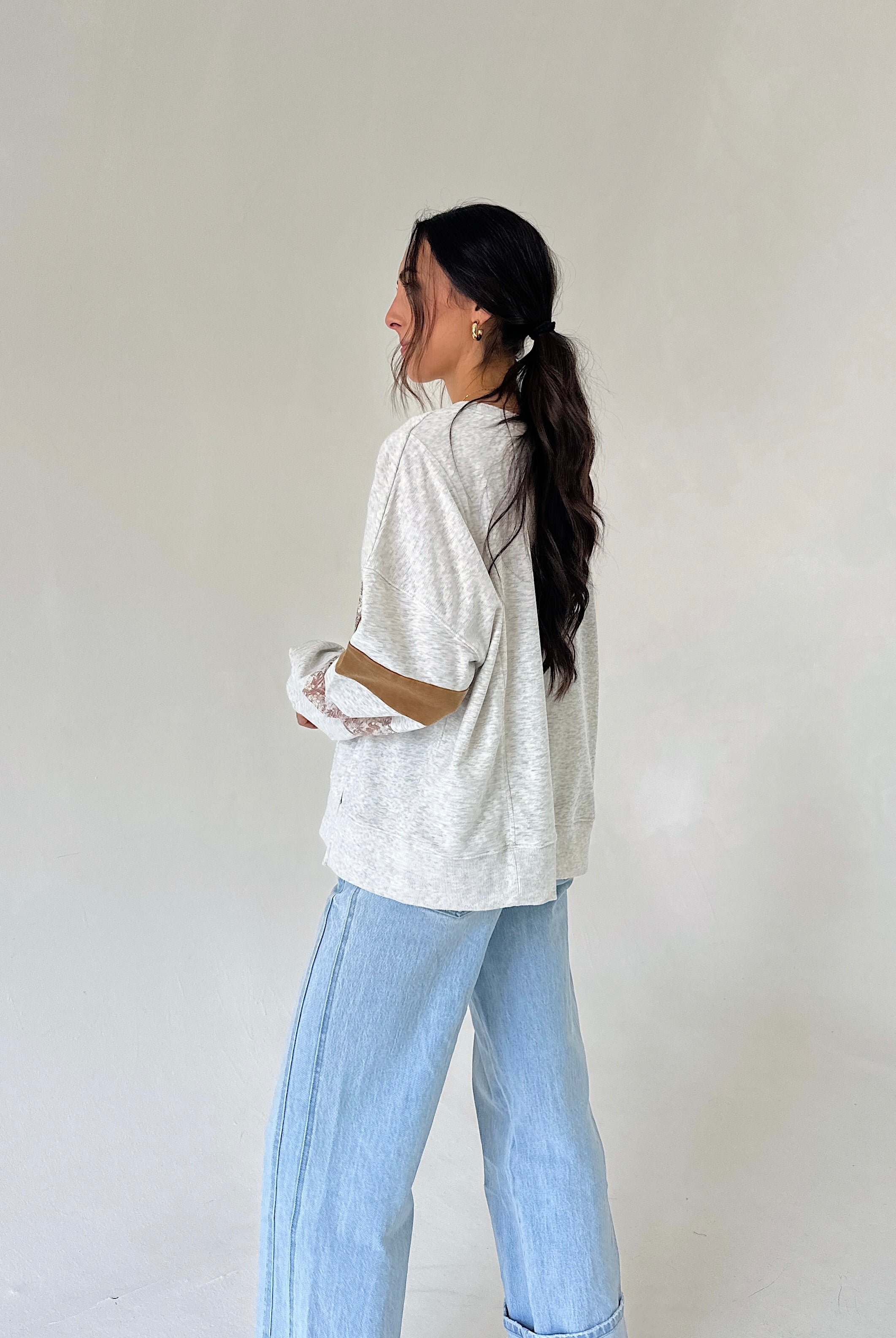 A woman with long dark hair tied back is shown from the side, wearing a heather grey long sleeve top and light blue wide-leg jeans against a plain off-white background.