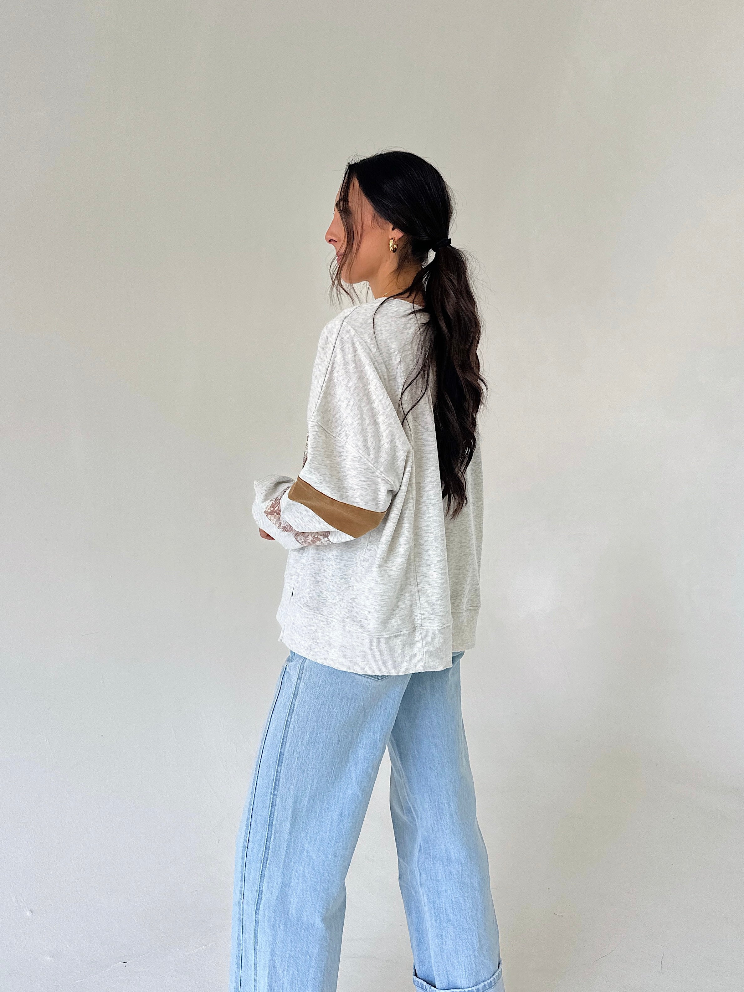 A woman with long dark hair tied back is shown from the side, wearing a heather grey long sleeve top and light blue wide-leg jeans against a plain off-white background.
