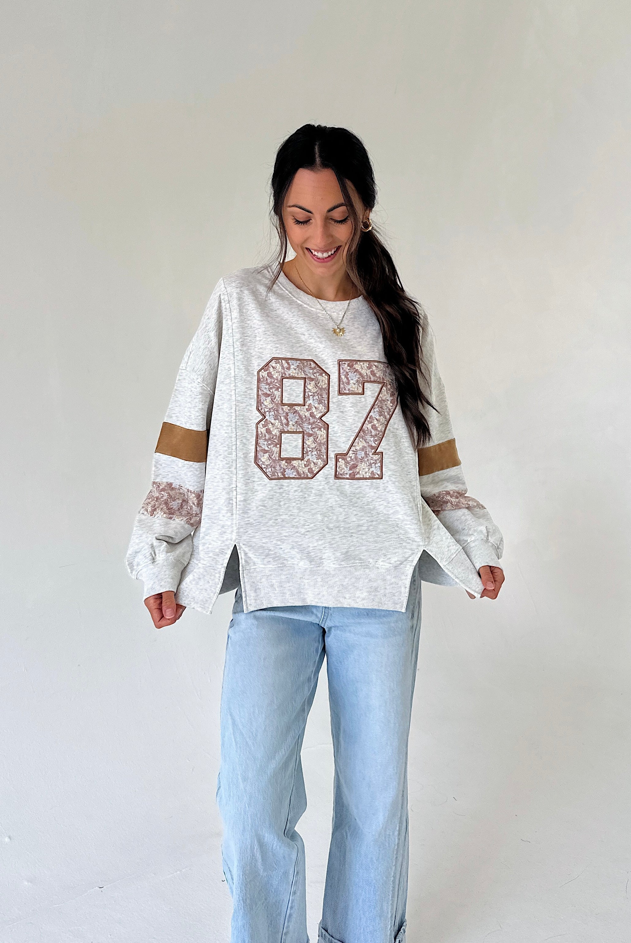 A woman modeling a white heathered long-sleeve sweatshirt with a large '87' graphic on the front, featuring lace-like detailing on the sleeves and light blue jeans, set against a plain off-white studio background.