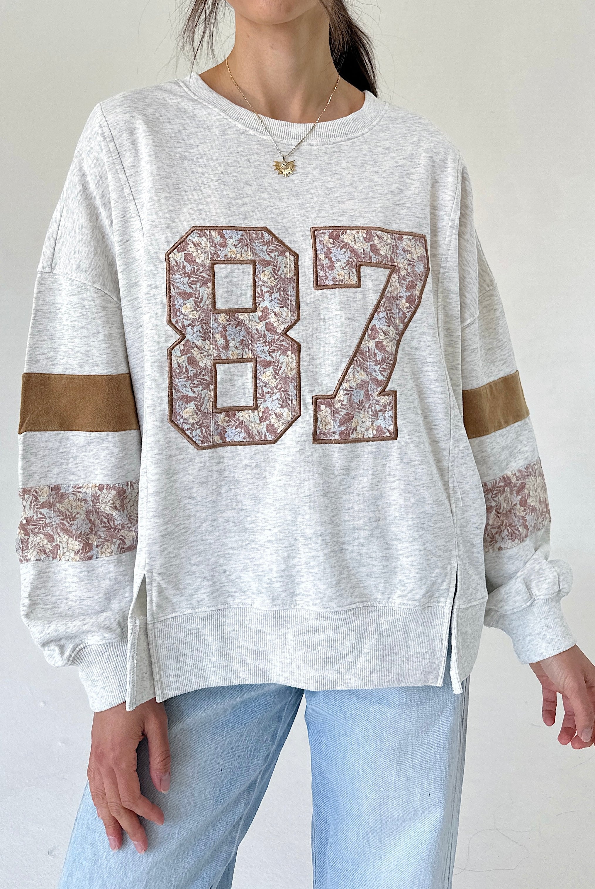 A woman wearing a light heather gray sweatshirt with a large floral-patterned '87' on the front, brown sleeve stripes, and light blue jeans, shown from the neck down against a plain light background.