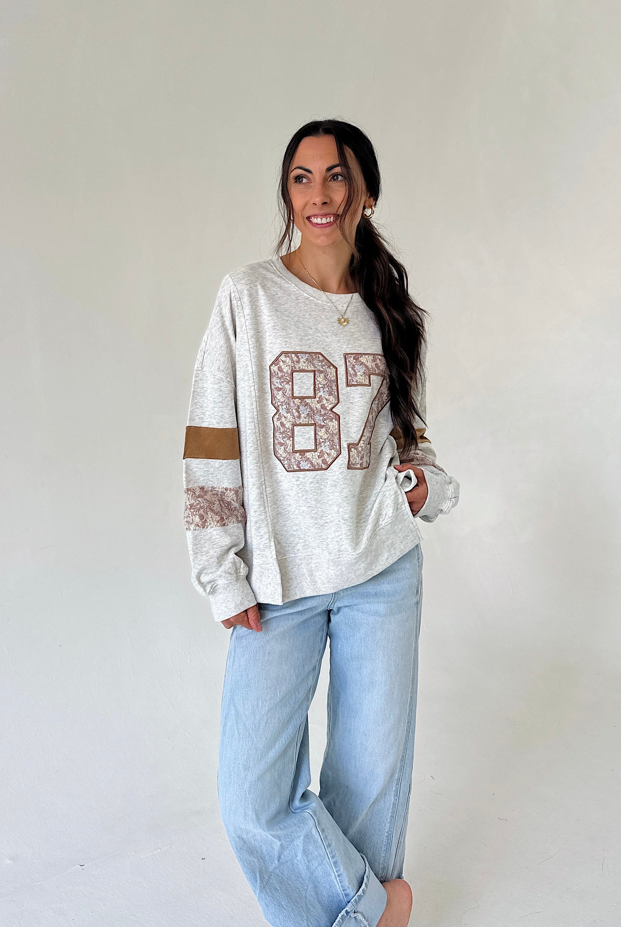 A woman modeling a light heather gray long-sleeve sweatshirt with a distressed brown and beige '87' graphic on the front, paired with light-wash blue jeans, against a plain off-white studio background.