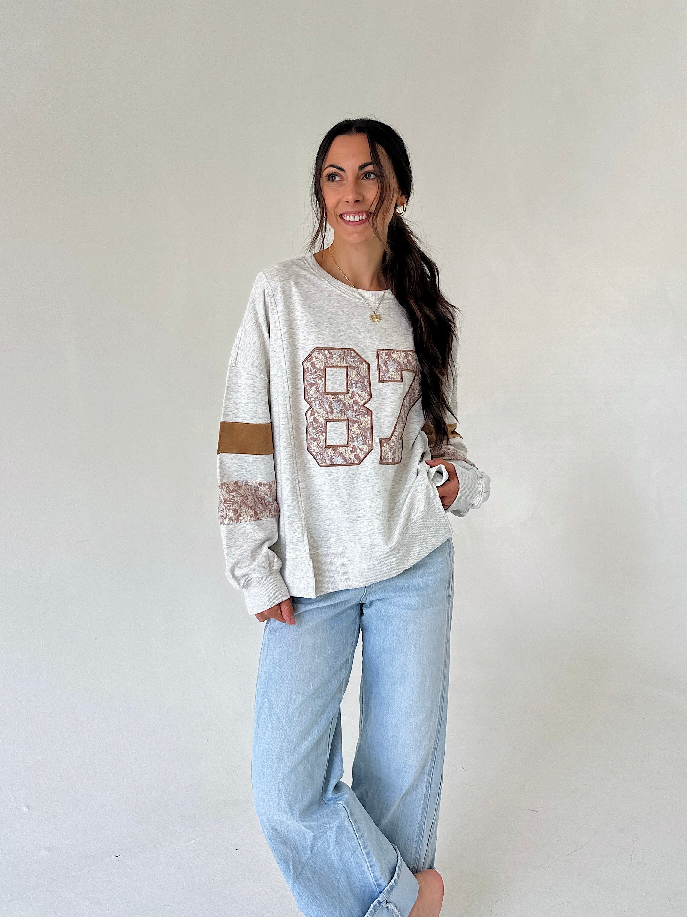 A woman modeling a light heather gray long-sleeve sweatshirt with a distressed brown and beige '87' graphic on the front, paired with light-wash blue jeans, against a plain off-white studio background.