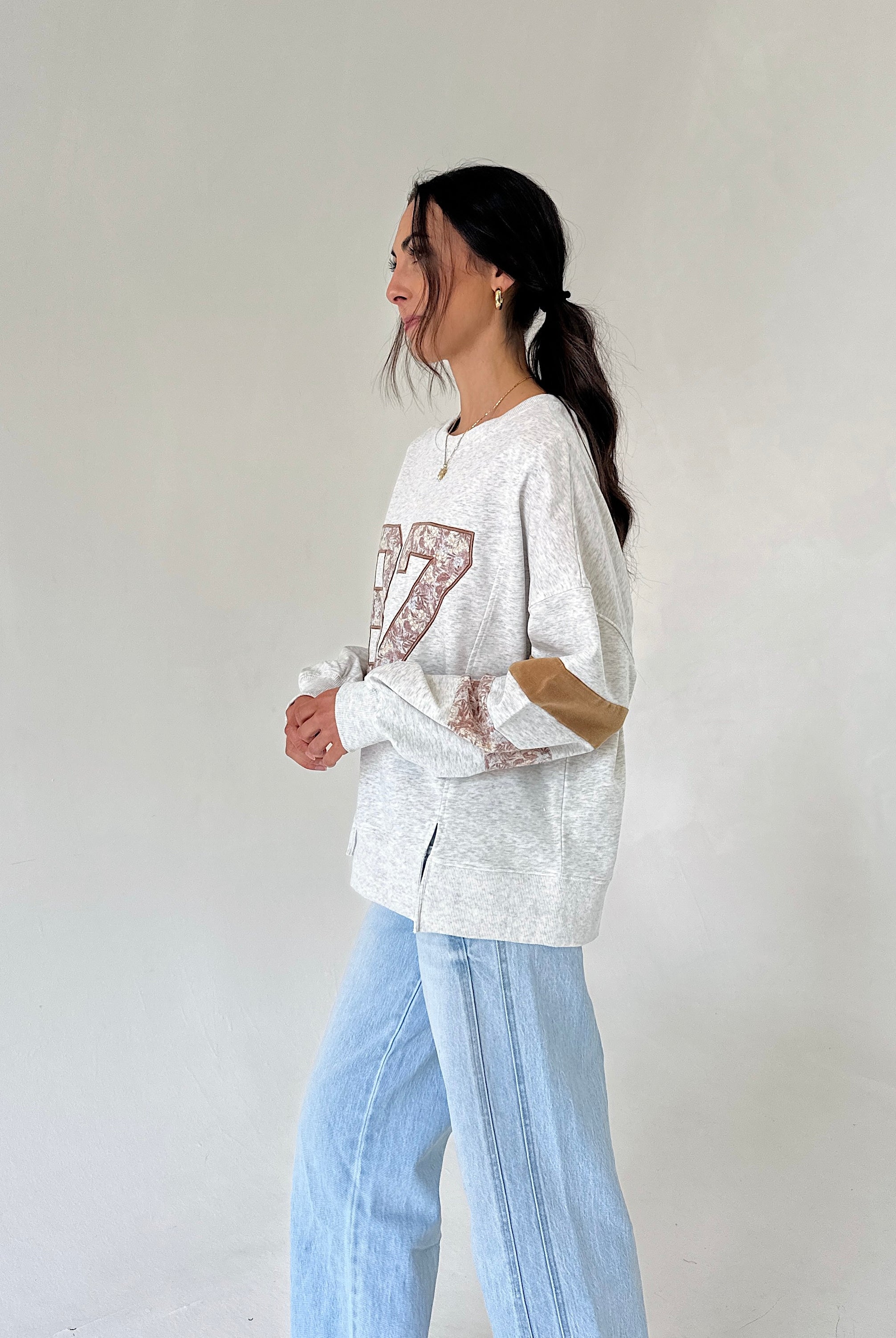 A woman wearing a light gray marled sweatshirt with a brown patterned graphic and light blue wide-leg jeans, standing against a plain off-white wall.