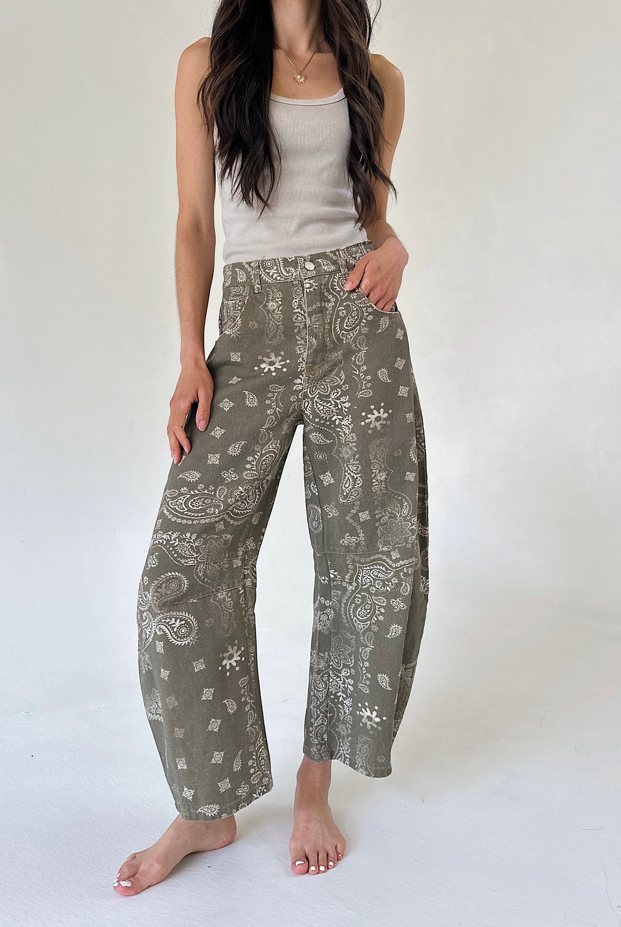 A woman modeling wide-leg barrel pants in an olive green paisley and floral print, paired with a white ribbed tank top, standing against a plain white studio background.