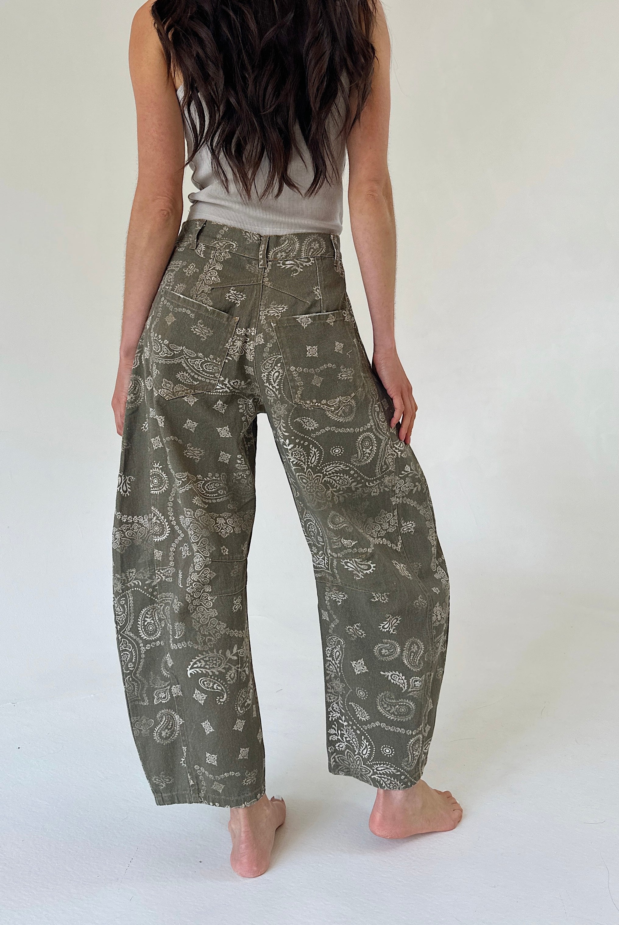 A person shown from the back wearing loose, wide-leg pants with an olive green and white paisley pattern, paired with a light gray sleeveless top, standing barefoot against a plain white studio background.