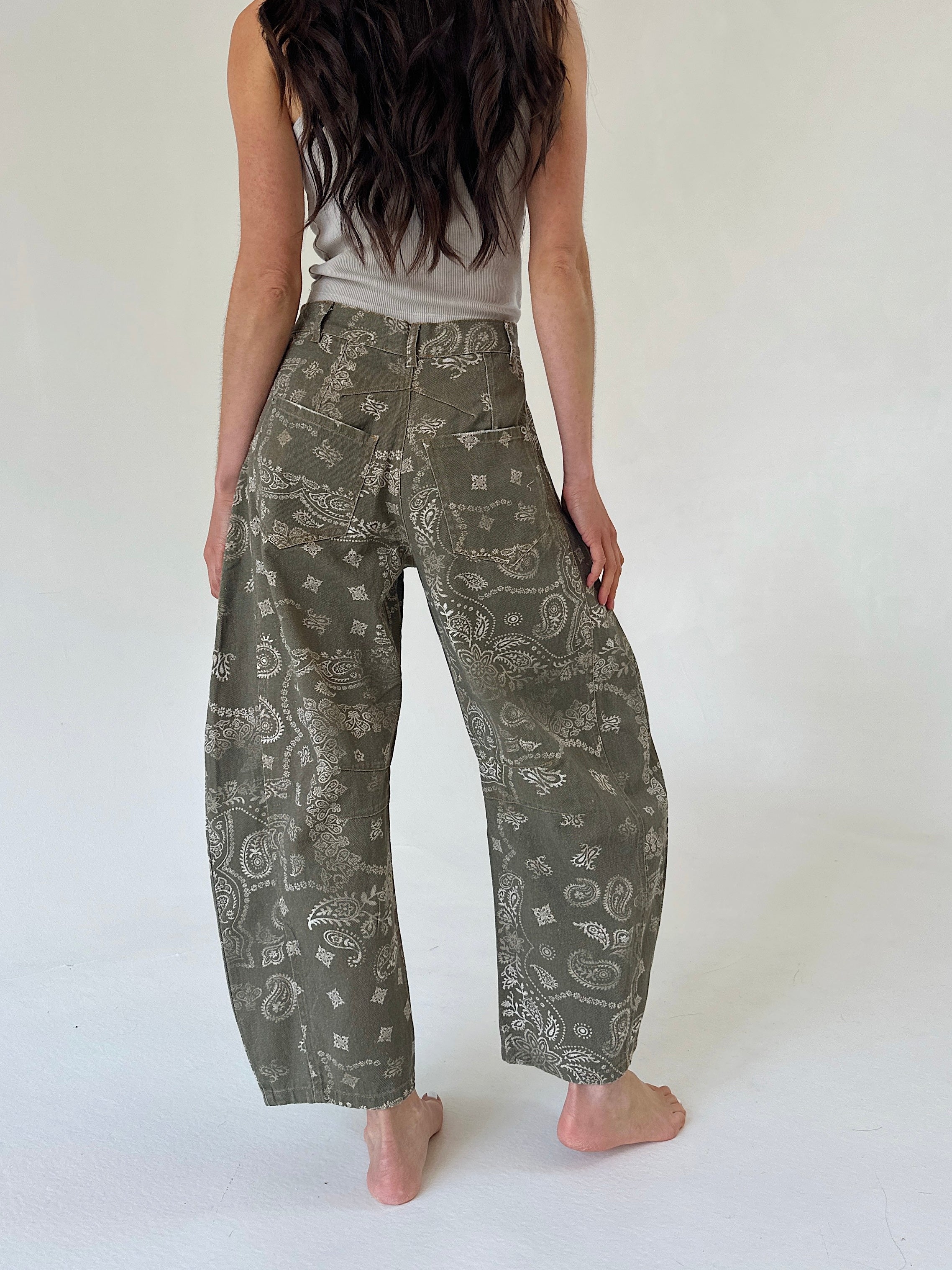 A person shown from the back wearing loose, wide-leg pants with an olive green and white paisley pattern, paired with a light gray sleeveless top, standing barefoot against a plain white studio background.