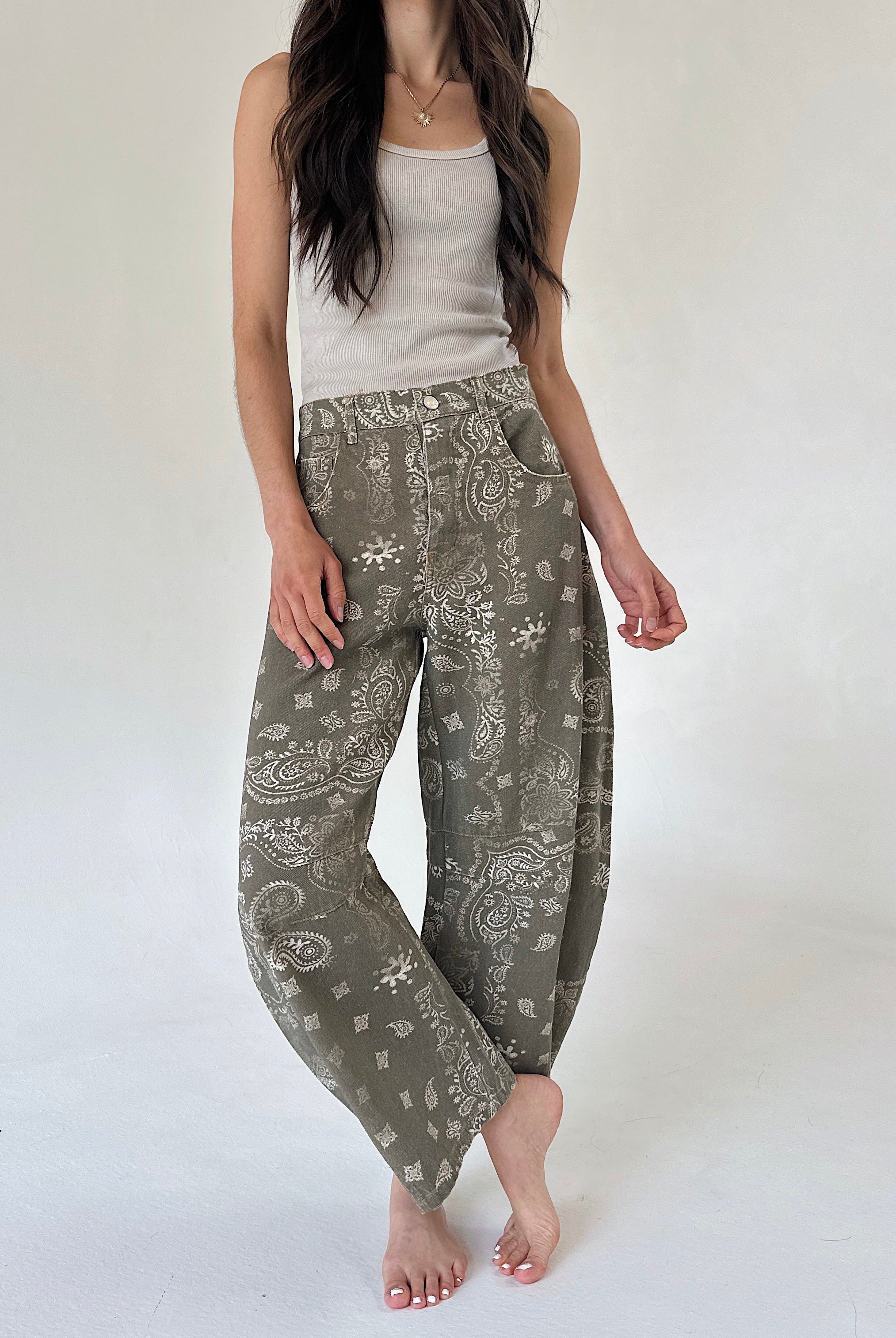 A woman modeling a pair of loose, wide-leg pants in an olive green paisley pattern, paired with a light beige tank top, against a plain off-white studio background.
