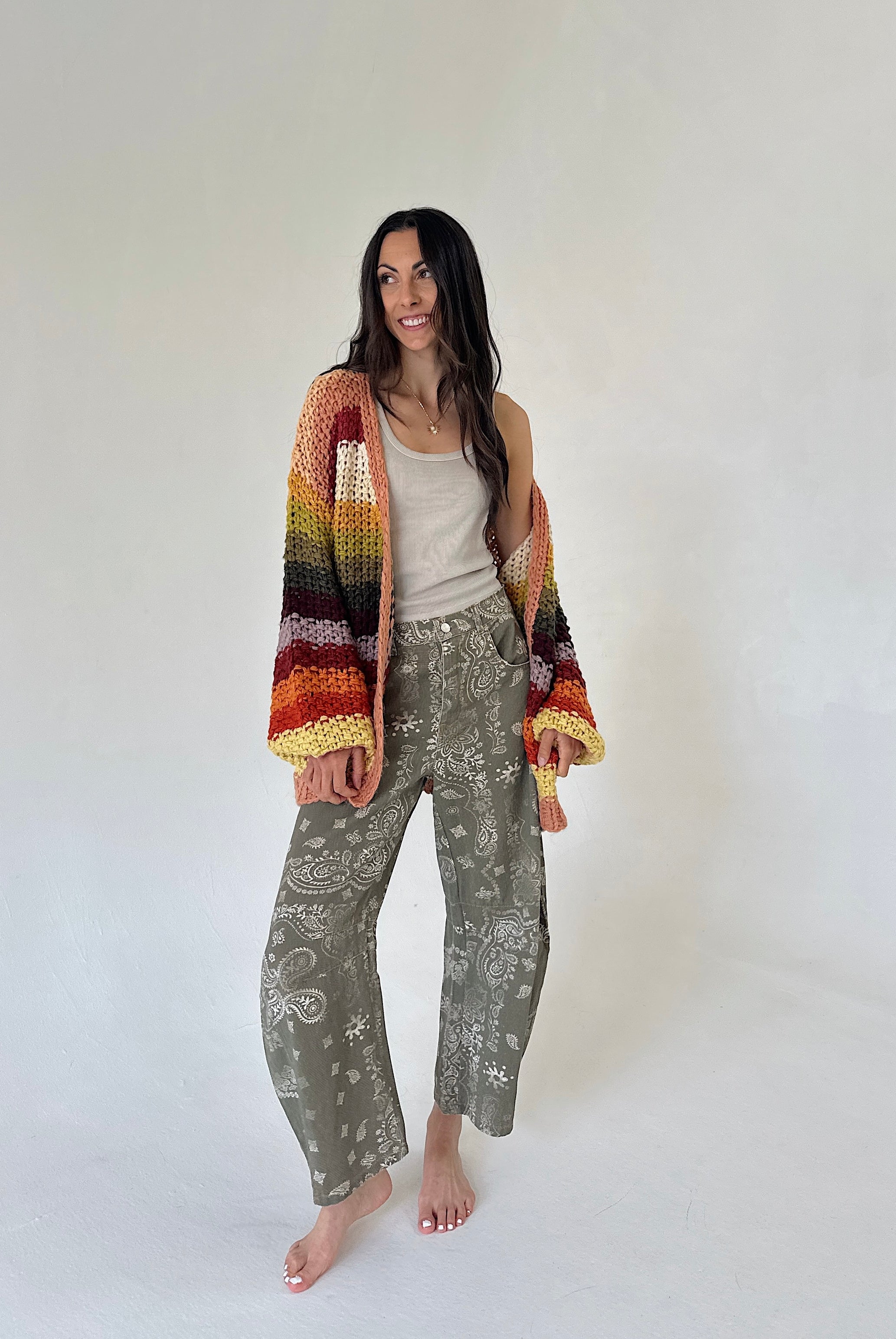 A woman stands barefoot against a plain light gray studio background, wearing a multicolor striped crochet cardigan with wide sleeves over a beige tank top and olive green patterned wide-leg barrel pants.