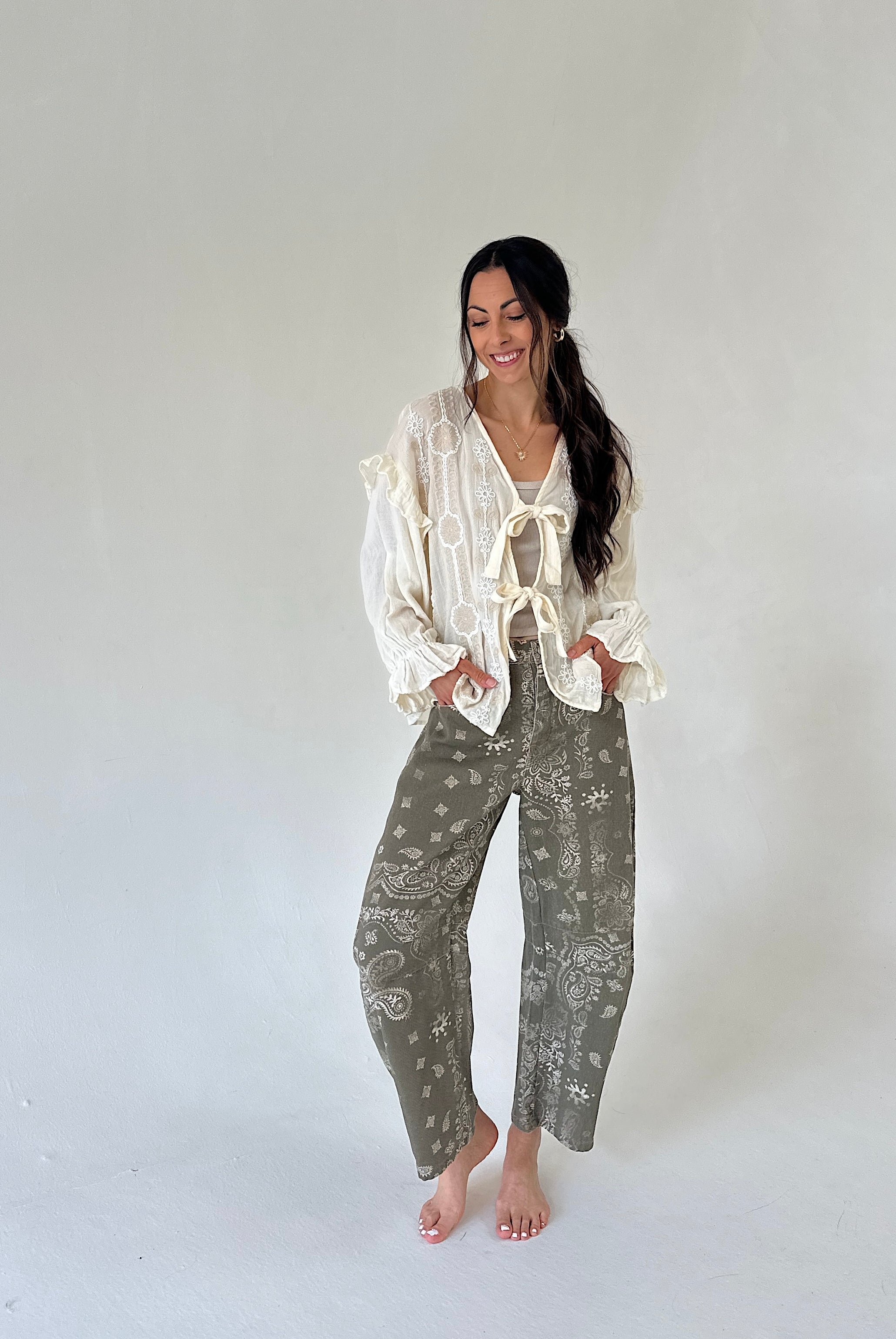 A woman modeling a bohemian-style outfit consisting of a cream lace blouse with ruffles and tie details, paired with olive green paisley bandana-print pants, standing against a plain light gray studio background.