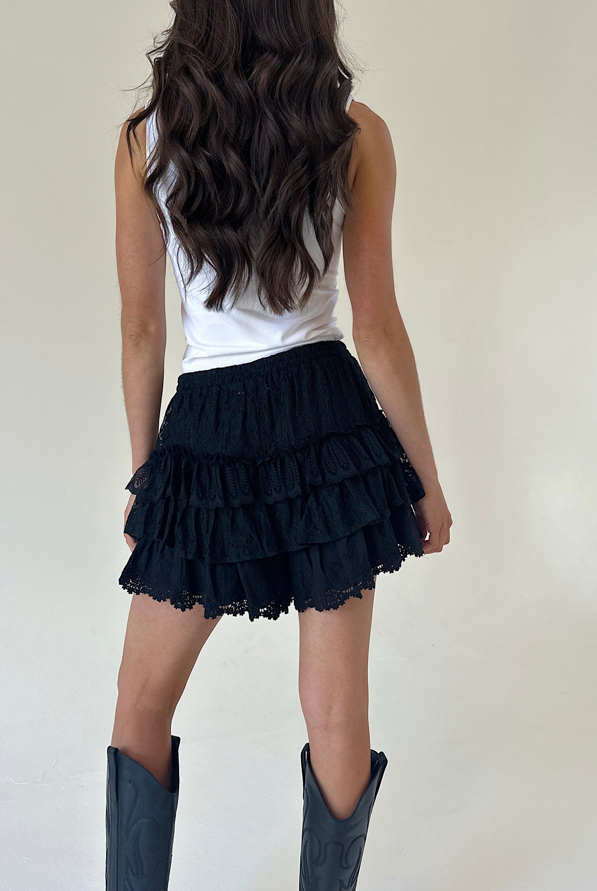 A woman shown from the back wearing black ruffled tiered shorts, a white sleeveless top, and dark cowboy boots against a plain off-white studio background.