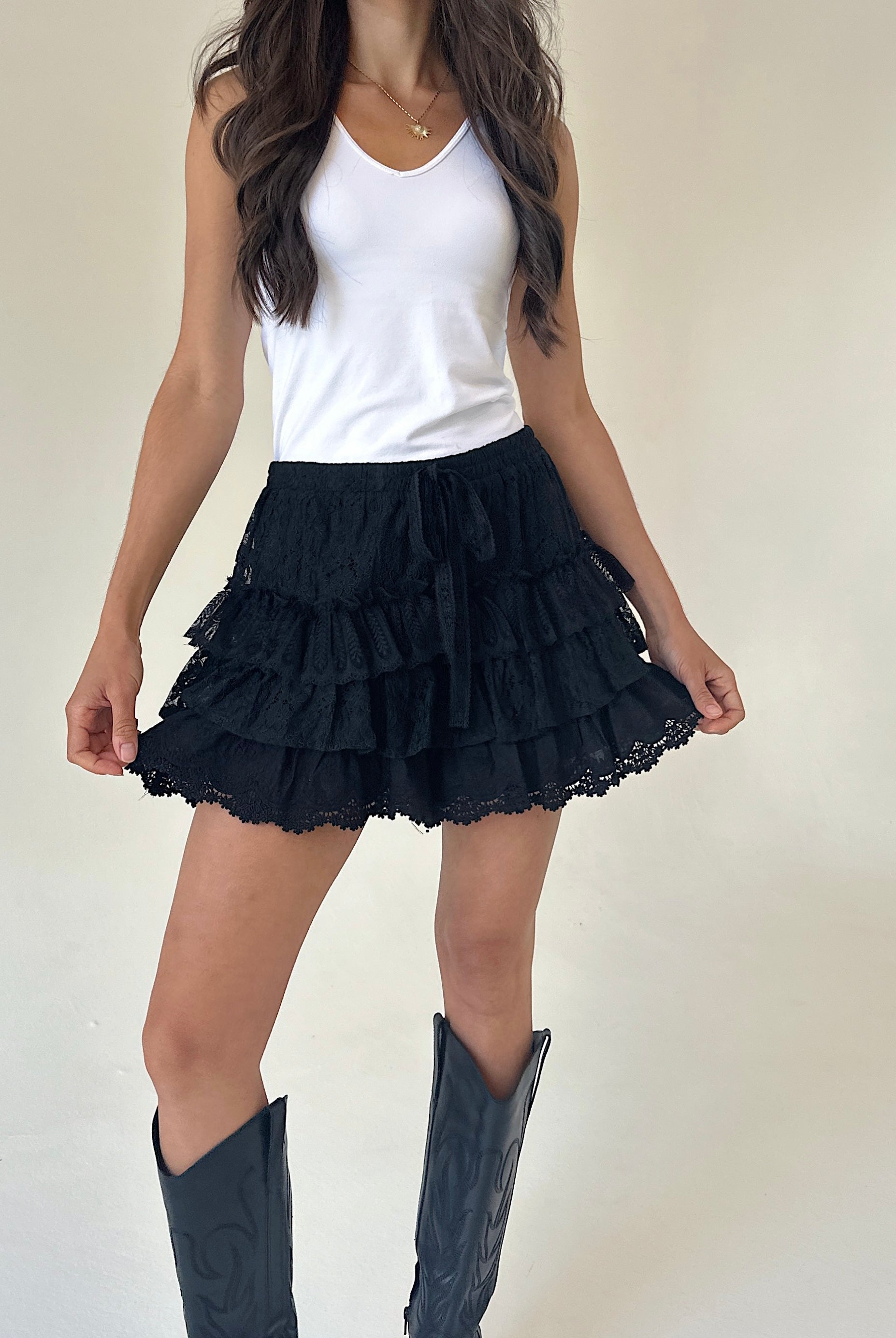A woman wearing black ruffled tiered shorts with lace trim, paired with a white tank top and black cowboy boots, standing against a plain off-white wall.