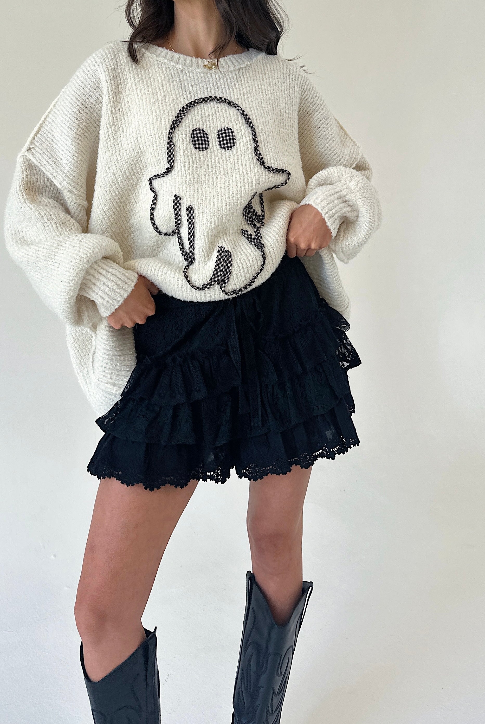A person wearing a cream knit sweater with a ghost motif, black ruffled shorts, and black cowboy boots, standing against a plain light background.