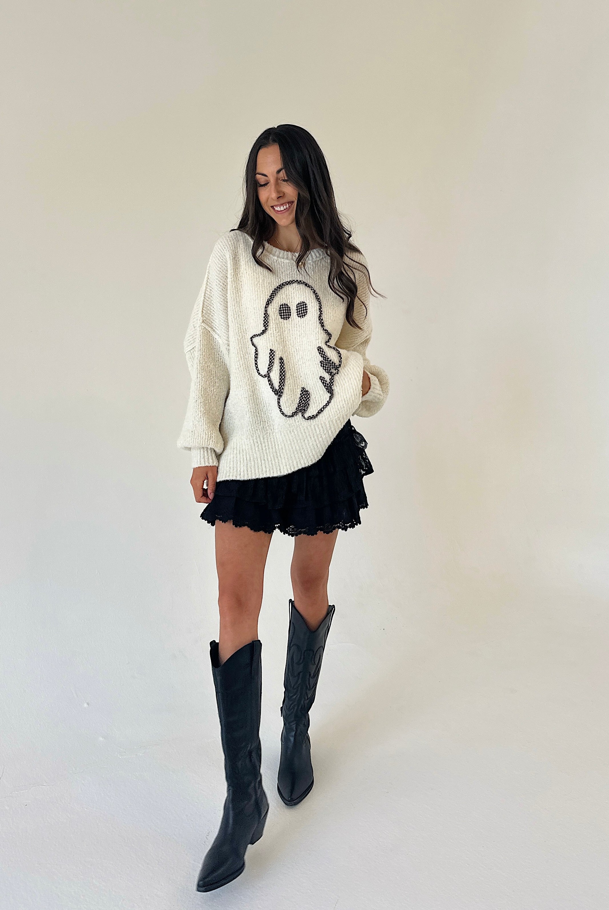 A woman modeling a cream knit sweater with a black ghost graphic, paired with a black ruffled mini short and black knee-high boots, standing against a plain off-white studio background.