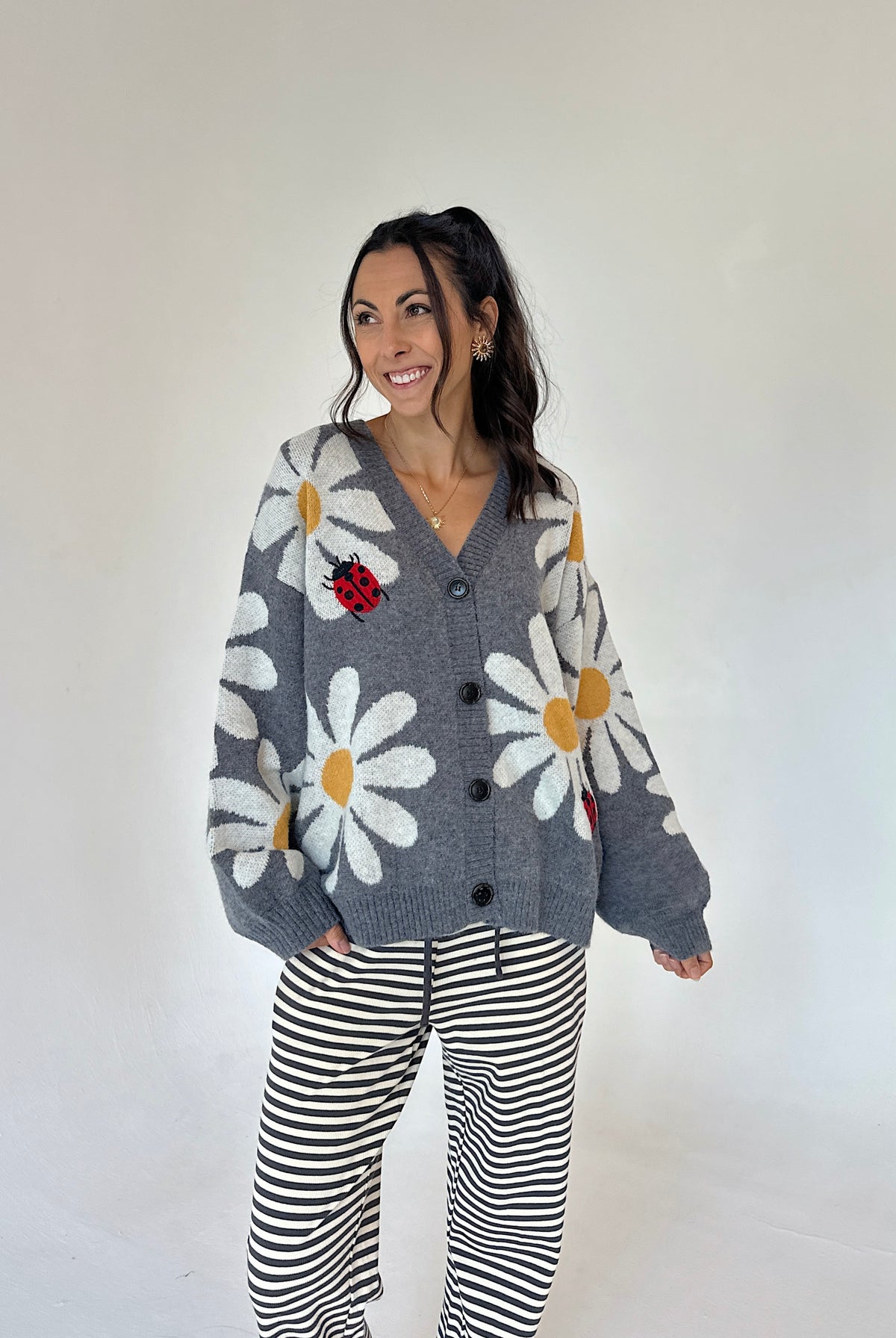 A woman modeling a gray knit cardigan with large white daisy patterns, yellow centers, and a red ladybug applique, paired with black and white striped pants, against a plain off-white studio background.
