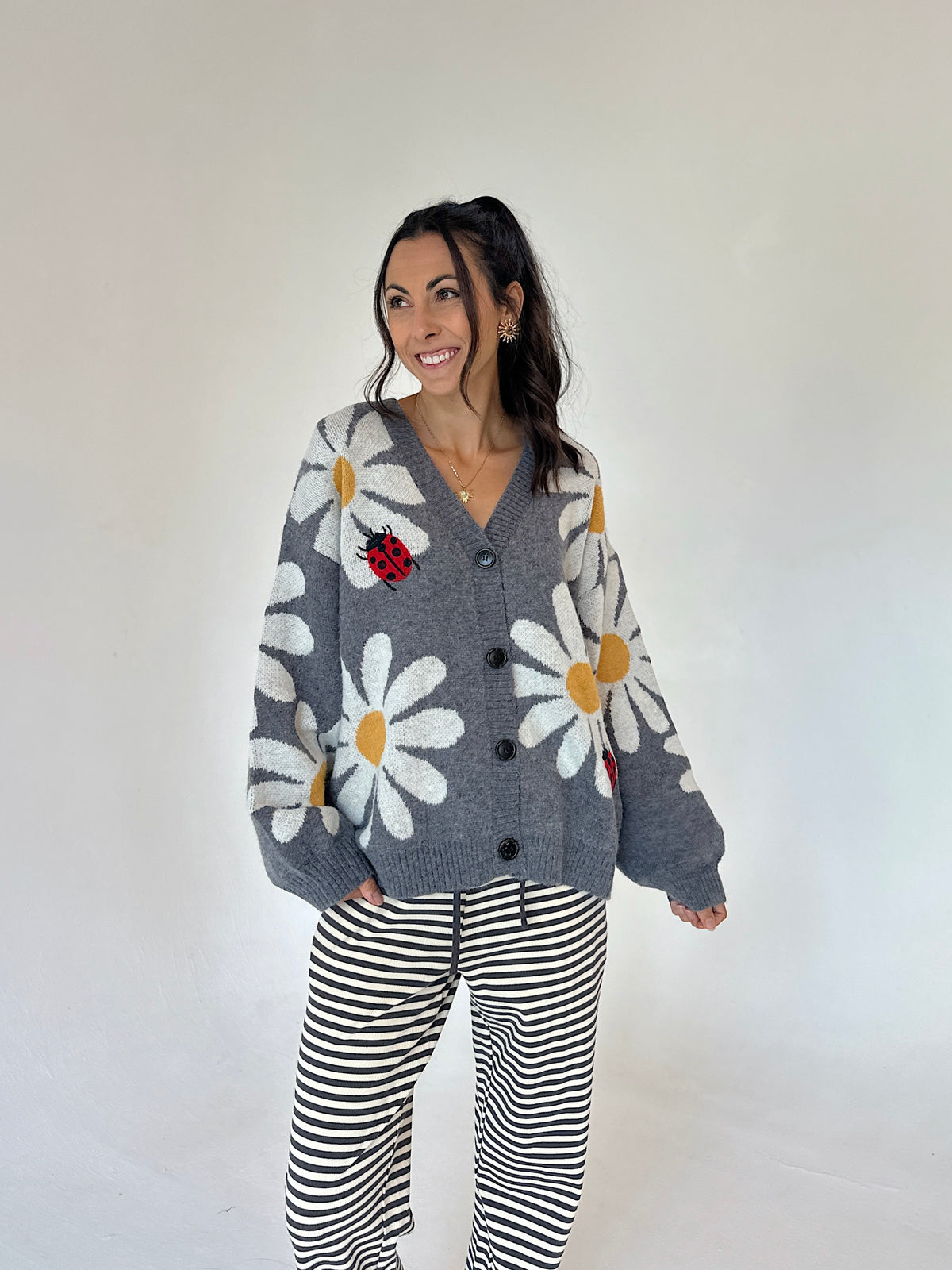A woman modeling a gray knit cardigan with large white daisy patterns, yellow centers, and a red ladybug applique, paired with black and white striped pants, against a plain off-white studio background.