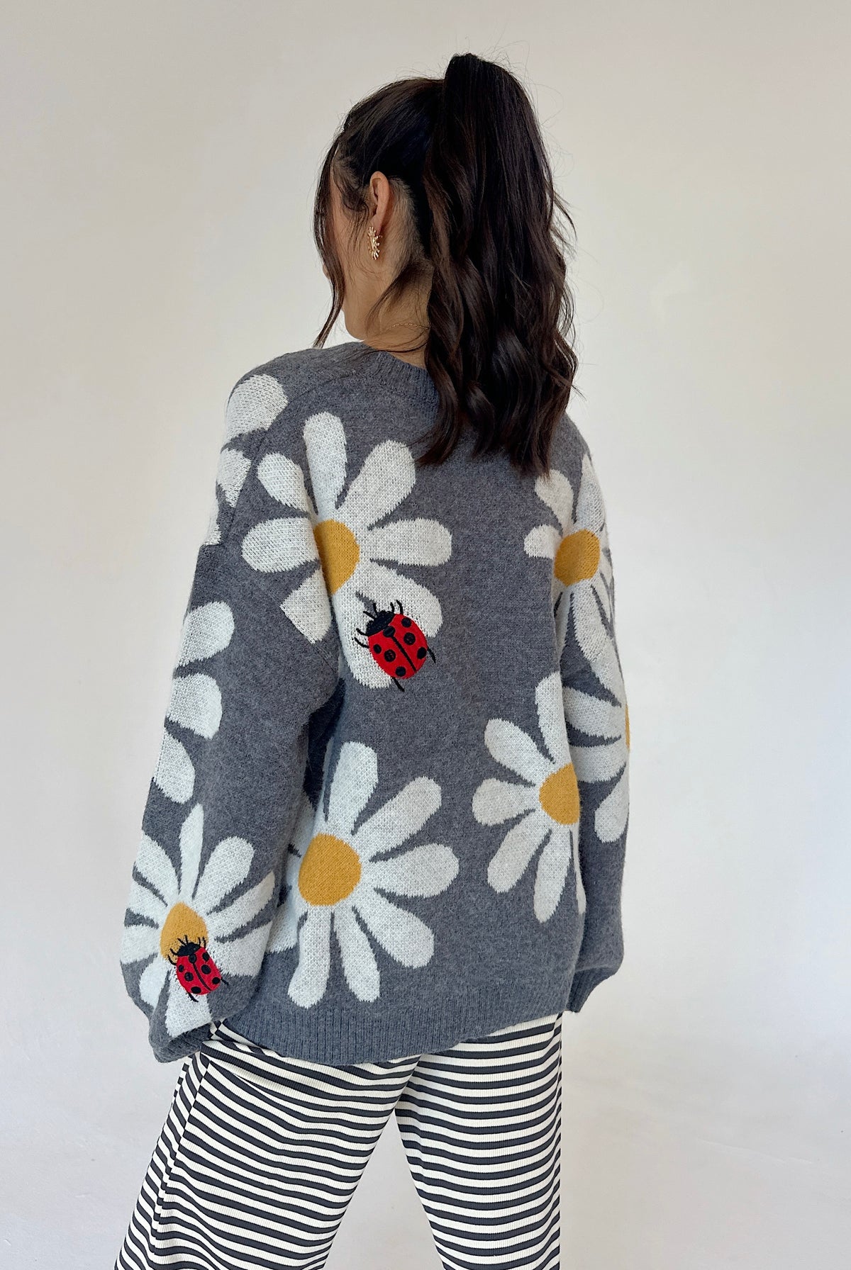 A woman viewed from behind wearing a gray knit sweater with large white daisy patterns, yellow centers, and red ladybugs, paired with black and white striped pants against a plain off-white background.