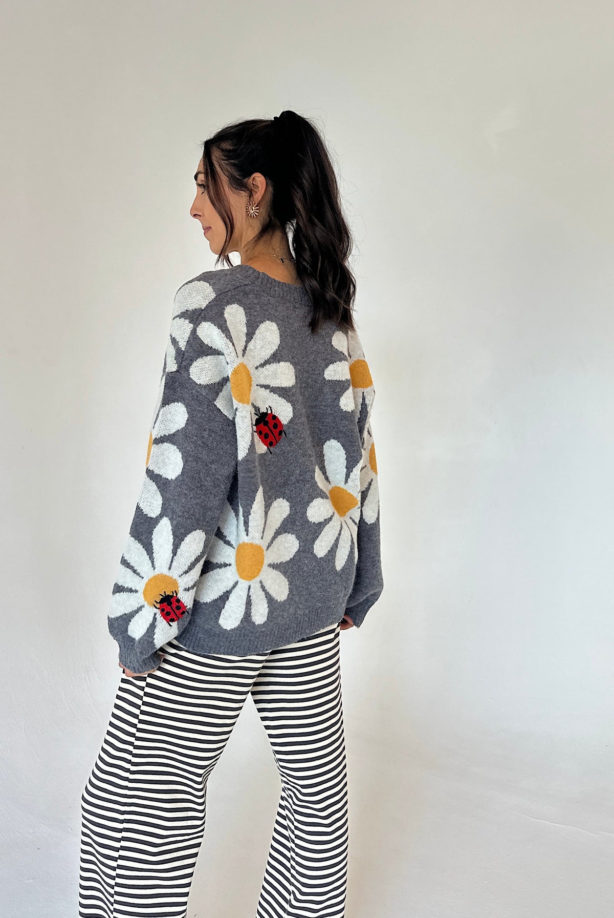 A woman is shown from the back wearing a gray knit sweater with large white daisy patterns, yellow centers, and small red ladybug appliqués, paired with black and white striped pants, against a plain off-white background.