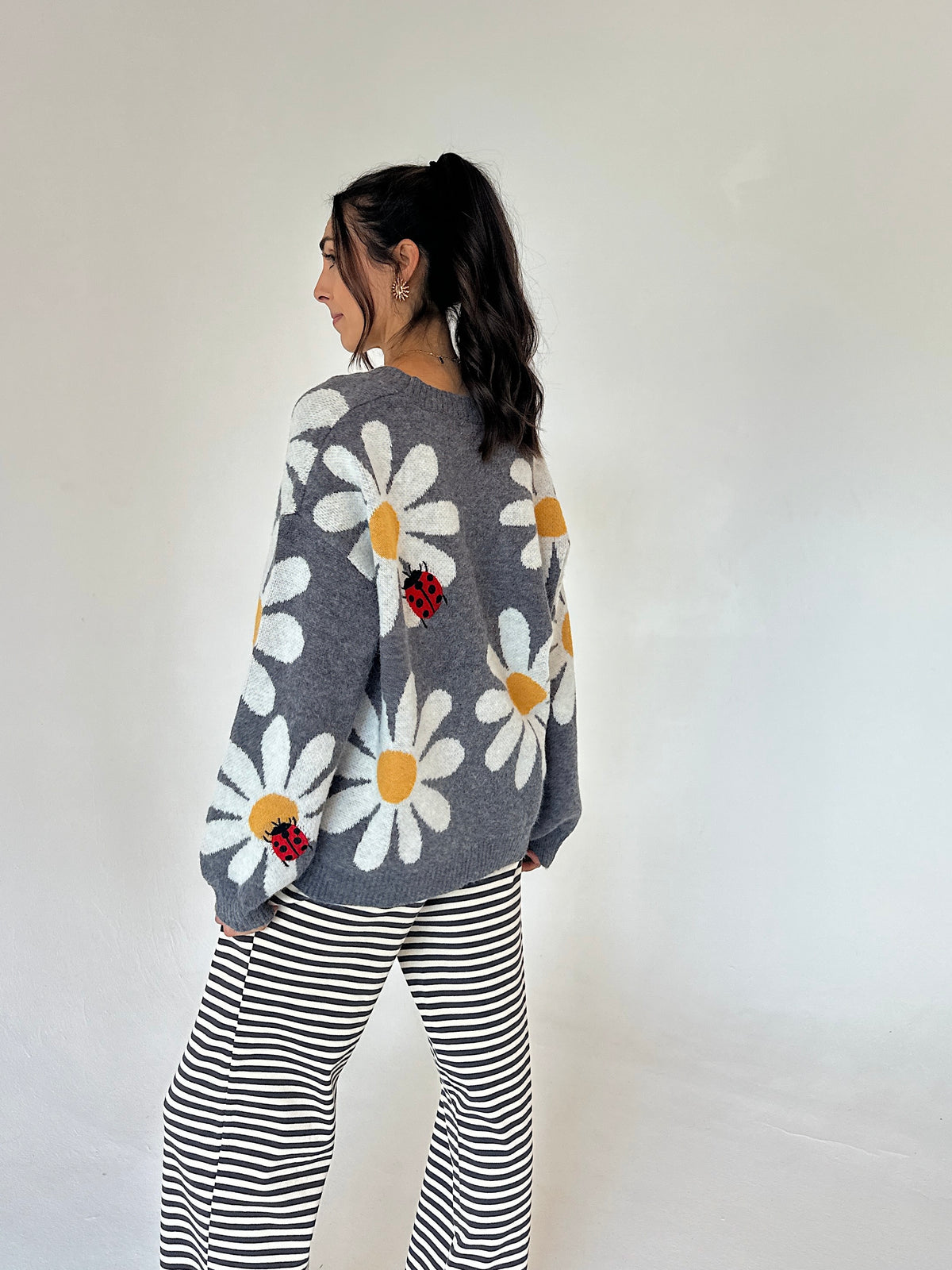 A woman is shown from the back wearing a gray knit sweater with large white daisy patterns, yellow centers, and small red ladybug appliqués, paired with black and white striped pants, against a plain off-white background.