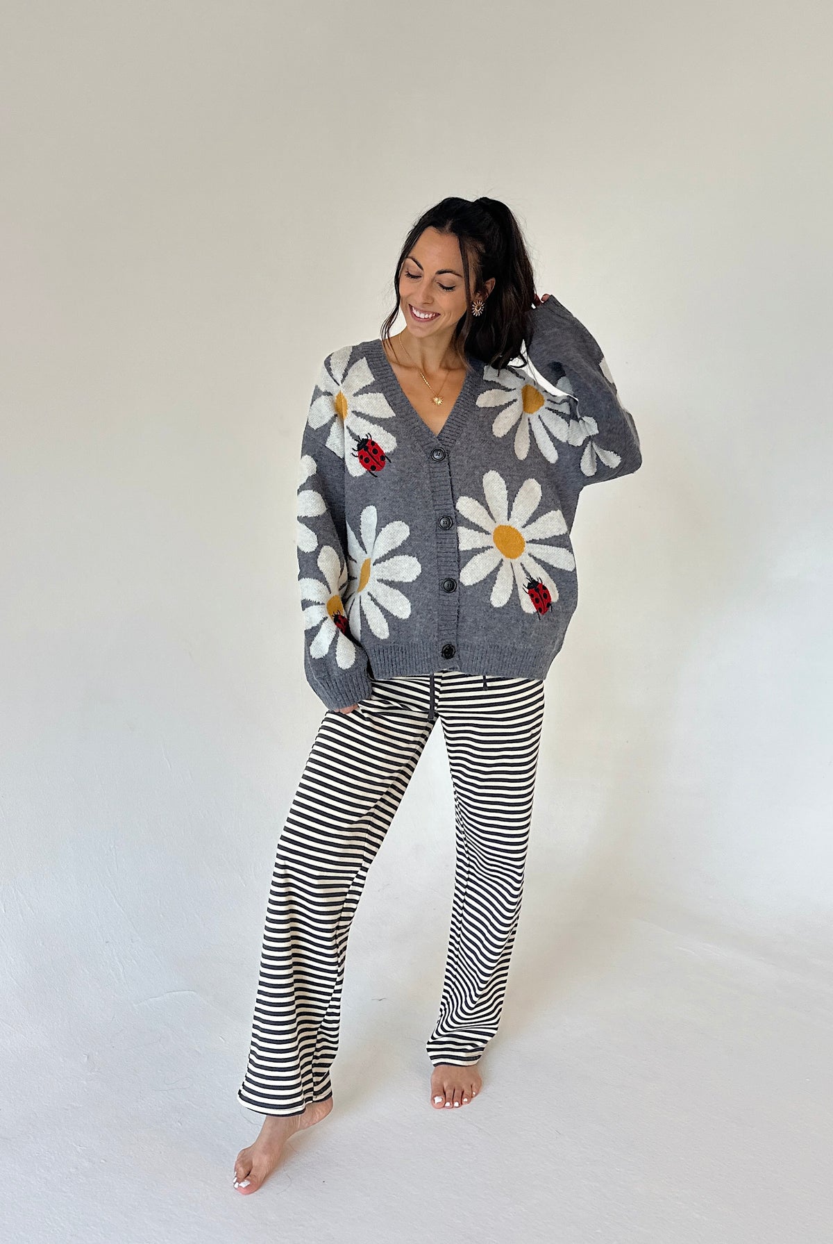 A woman modeling a cozy gray knit cardigan with large white daisy and ladybug motifs, paired with black and white horizontally striped pants, against a plain off-white studio background.