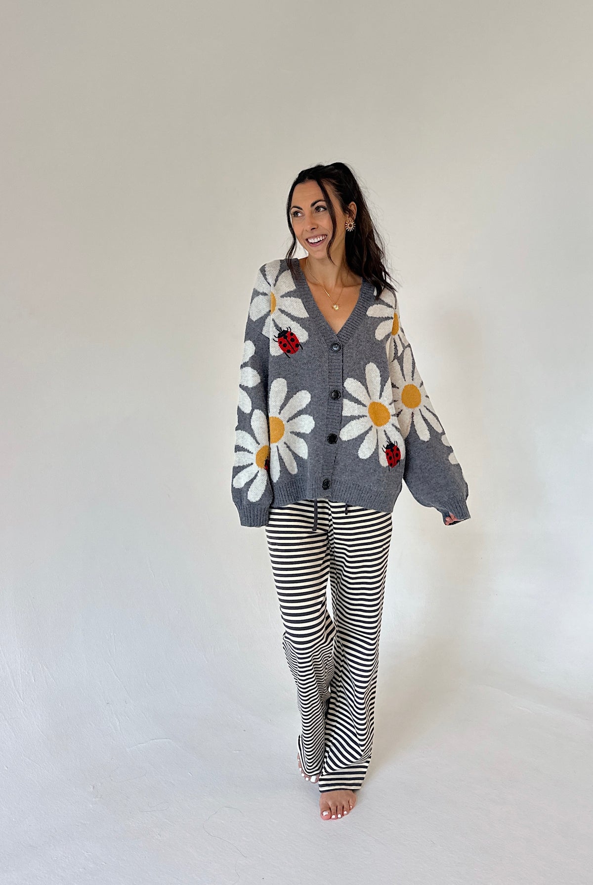 A woman modeling a gray knit cardigan with large white daisy patterns and red ladybug accents, paired with black and white striped pants, against a plain white studio background.