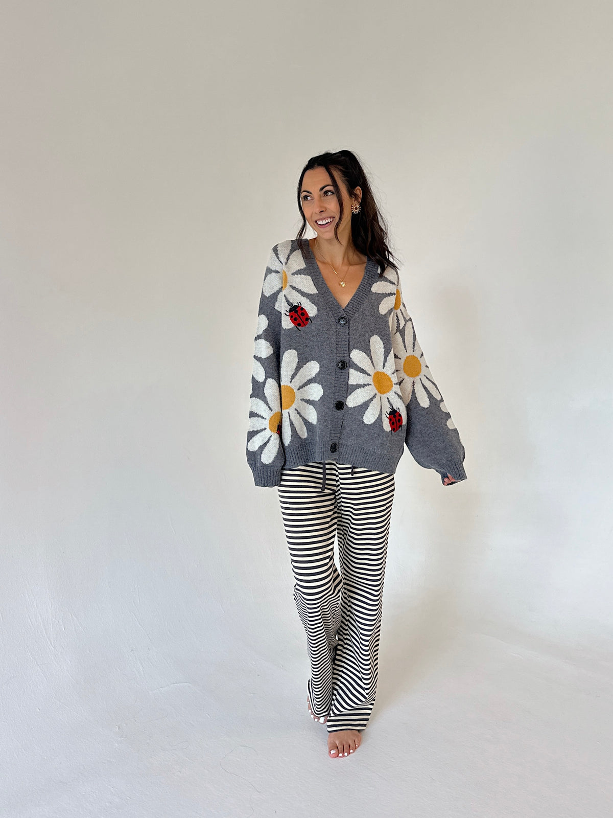 A woman modeling a gray knit cardigan with large white daisy patterns and red ladybug accents, paired with black and white striped pants, against a plain white studio background.