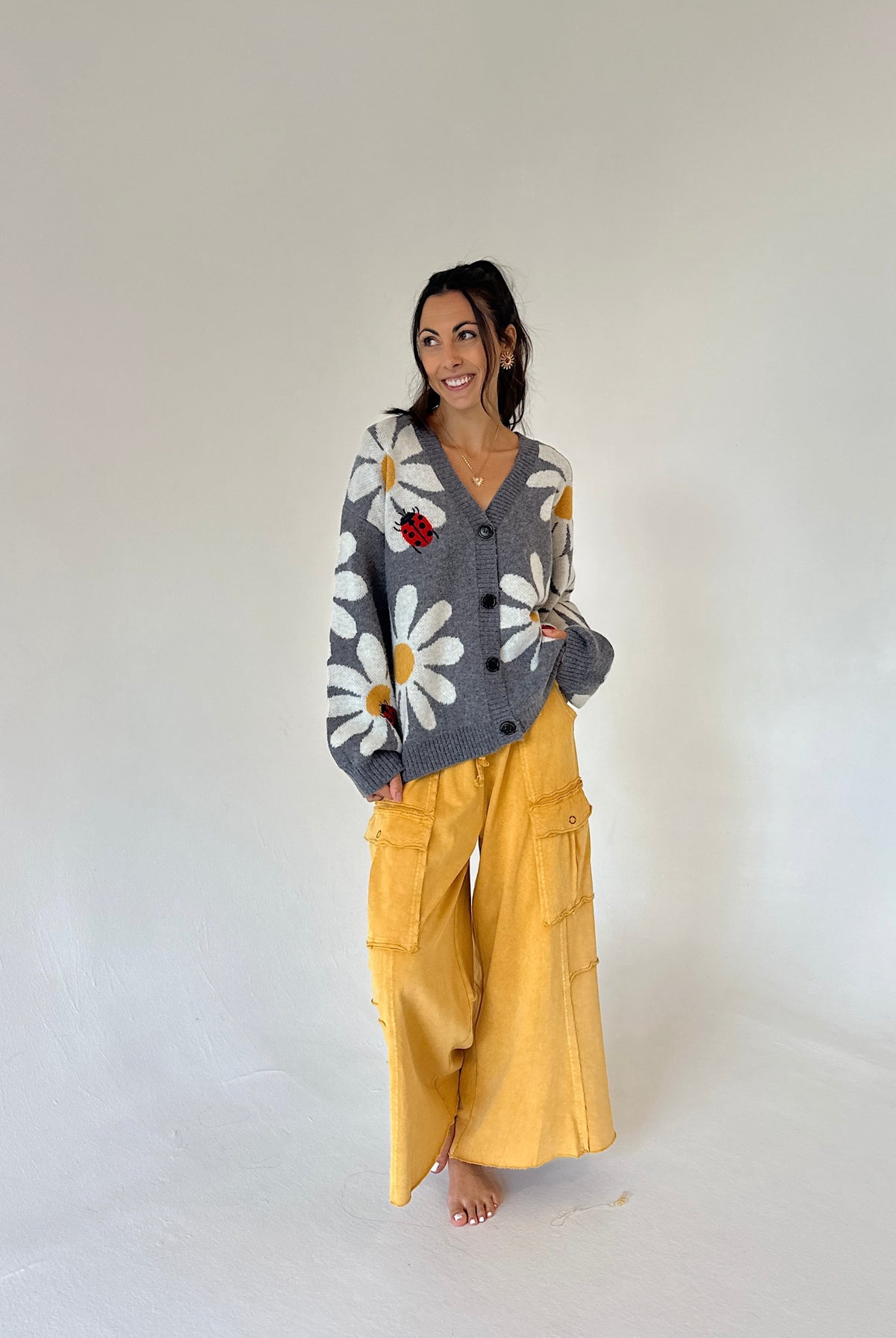 A woman stands barefoot against a plain light gray studio background, wearing a gray knit cardigan with large white daisy patterns and a small red flower accent, paired with wide-leg mustard yellow pants.