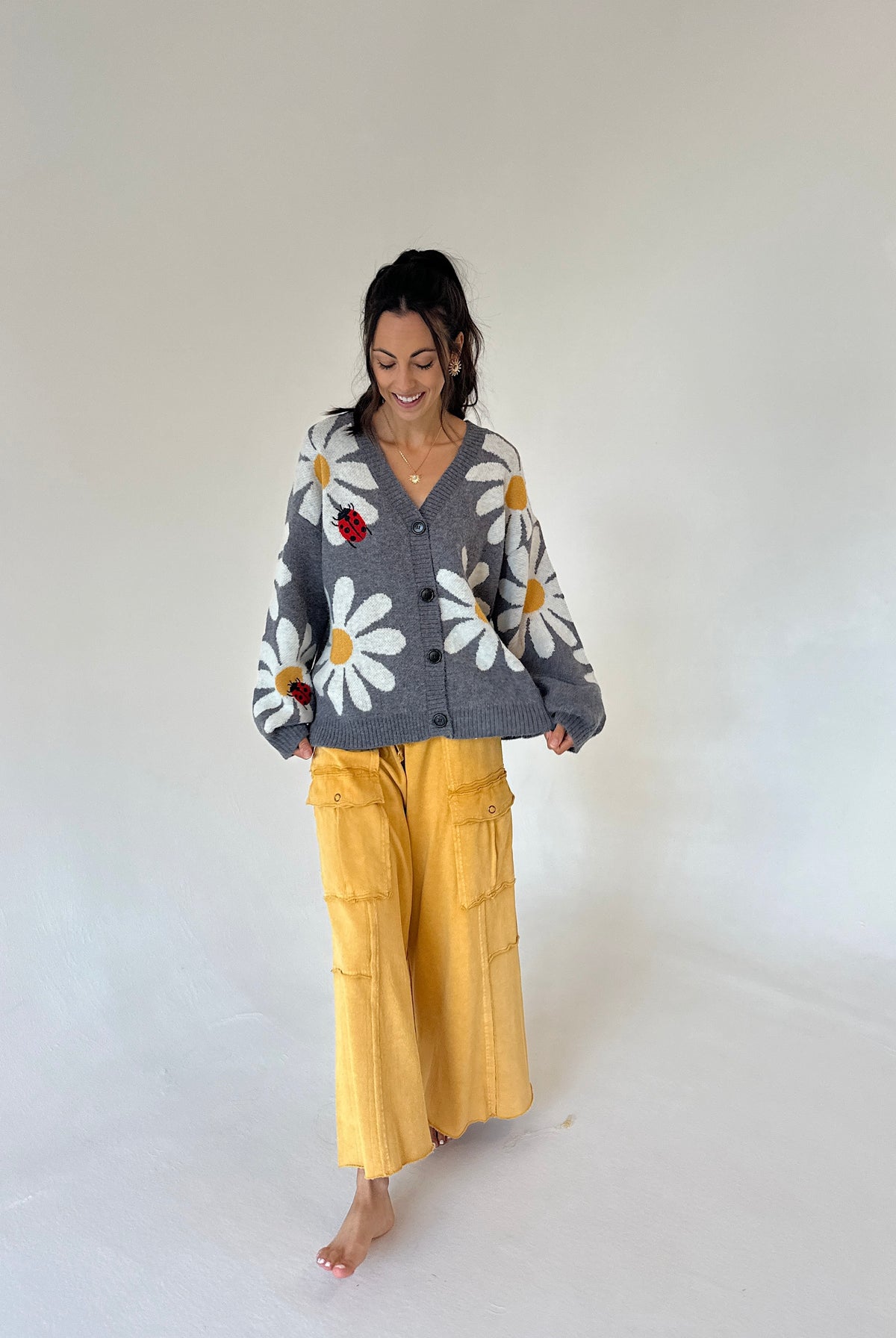 A woman stands barefoot against a plain white studio background, wearing a gray knit cardigan with large white daisy flowers and yellow cargo pants.
