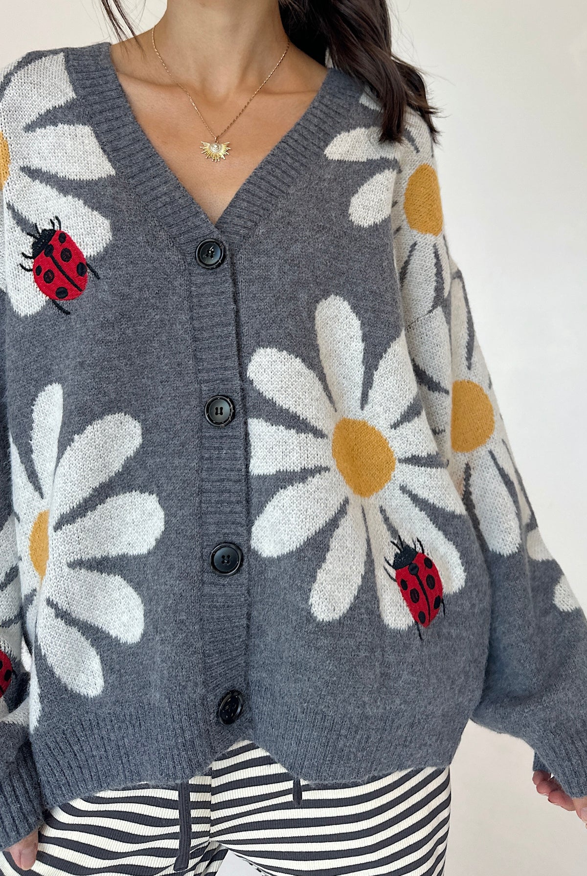 A woman wearing a gray knit cardigan with large white daisy patterns, yellow centers, and red ladybugs, paired with black-and-white striped pants.
